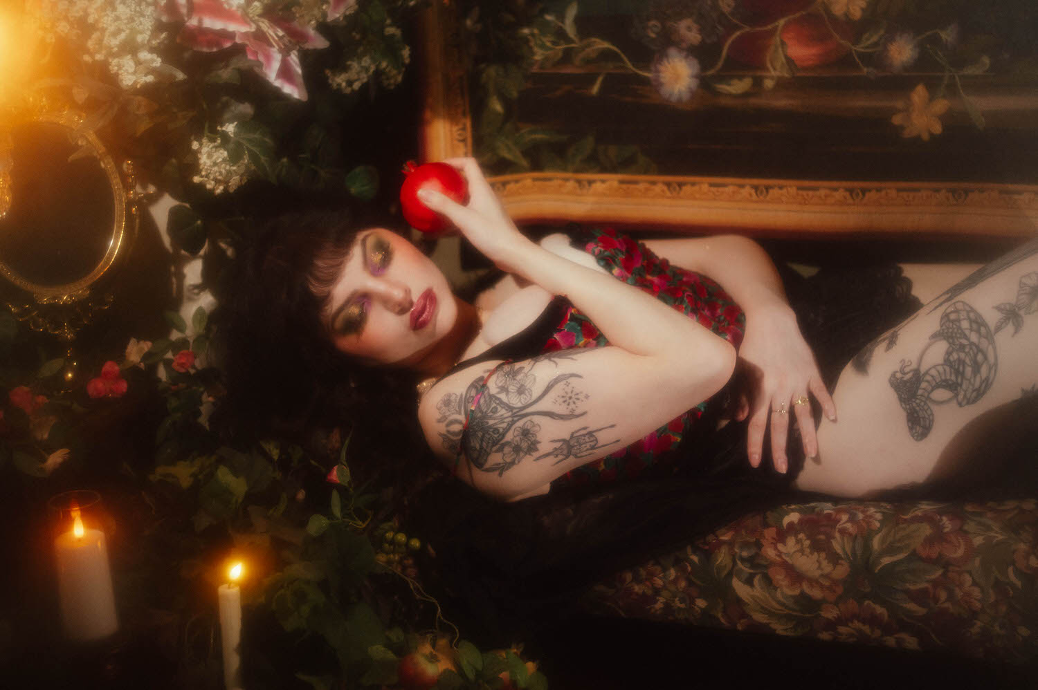 A woman reclines on a floral upholstered vintage chaise holding a red pomegranate, with visible arm and thigh tattoos and lit candles glowing nearby in this dark romantic boudoir photography session, Royal Lune Photo.