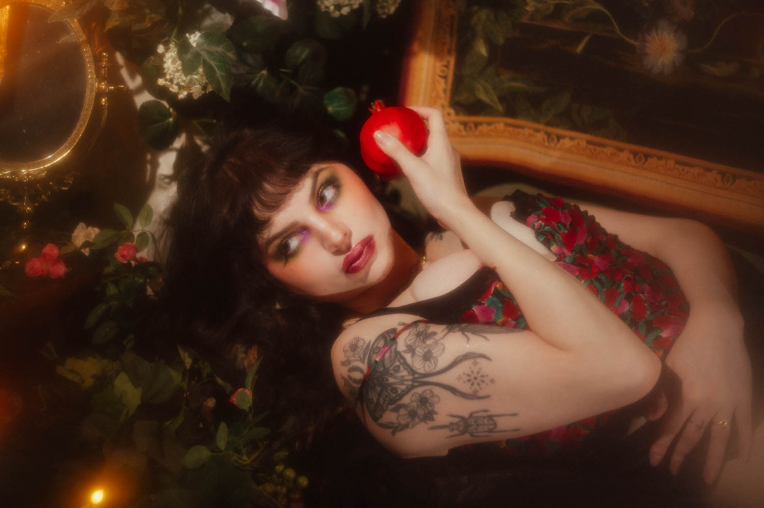 A woman in side profile holds a red pomegranate near her face, wearing a floral print lingerie top and layered gold necklaces beside a gold framed antique mirror in this vintage lingerie boudoir photoshoot, Royal Lune Photo.