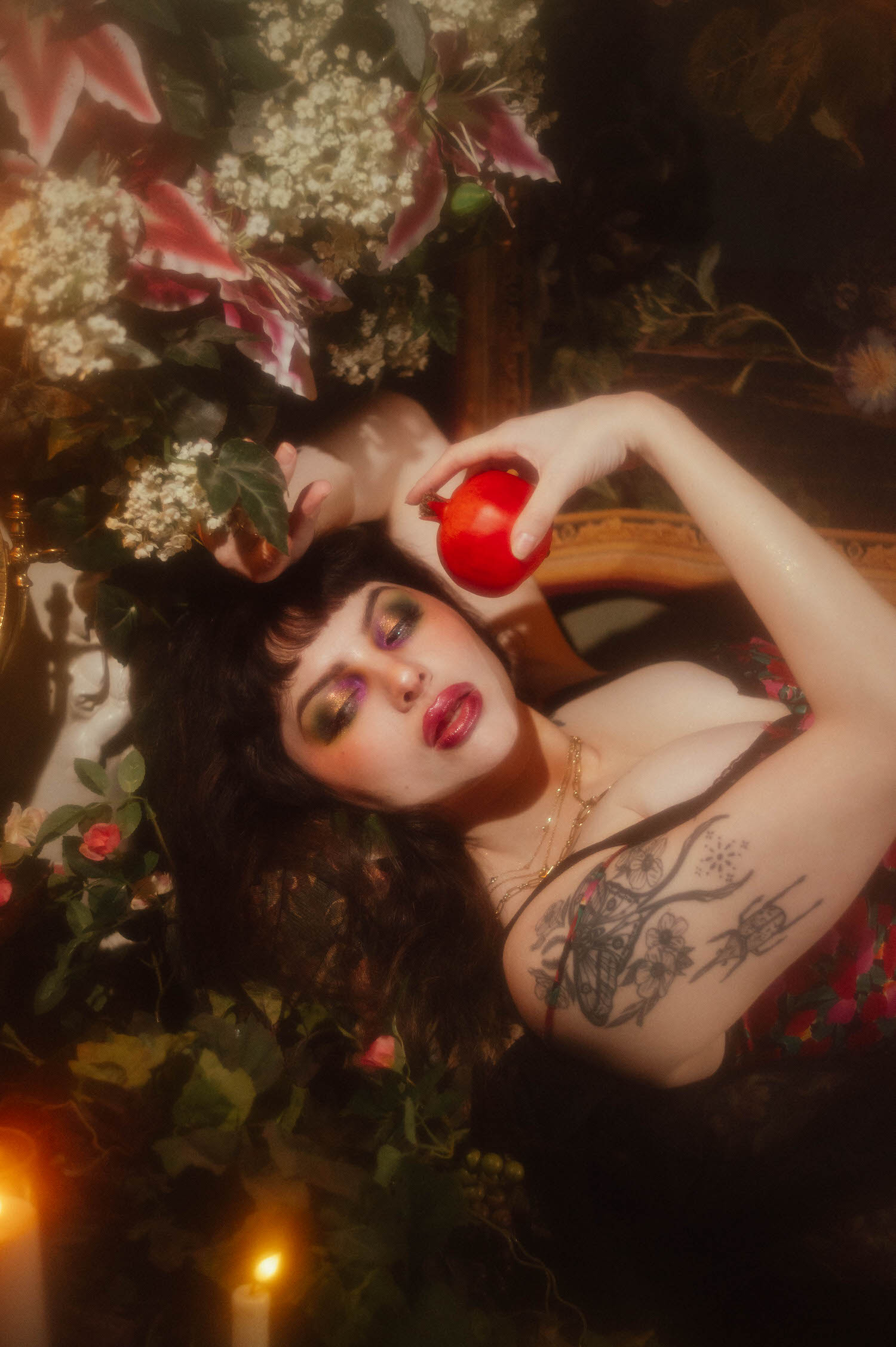 A woman lies with her eyes closed holding a red pomegranate above her, a floral tattoo visible along her ribcage, surrounded by pink lilies and baby's breath in this red rose boudoir photoshoot at Royal Lune Studio, Royal Lune Photo.