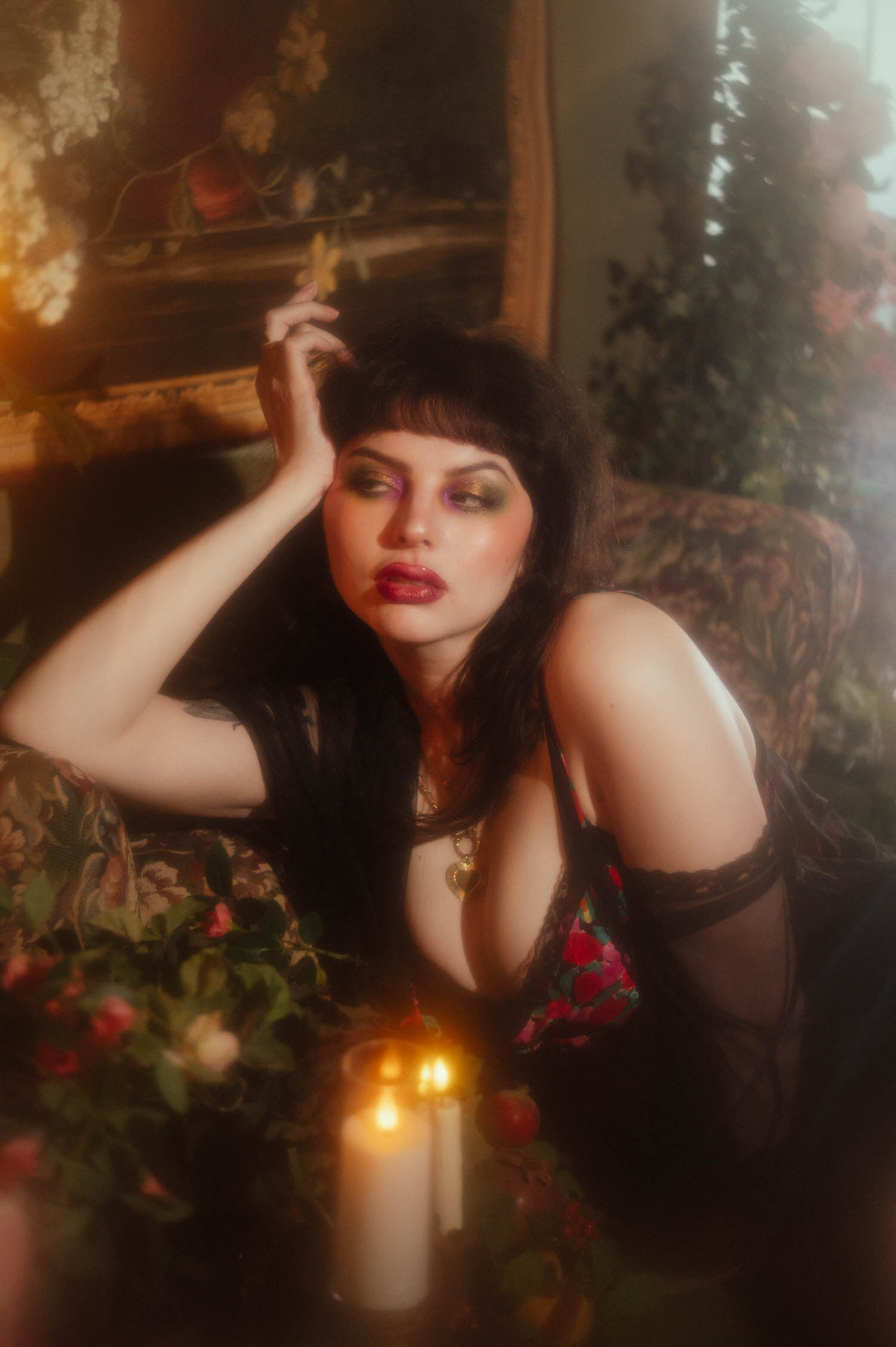 A woman leans forward resting her arm on a floral upholstered chair, wearing a black lace and floral print lingerie set with sheer black thigh highs, lit candles glowing in the foreground, a good example of boudoir photoshoot ideas built around vintage florals, Royal Lune Photo.