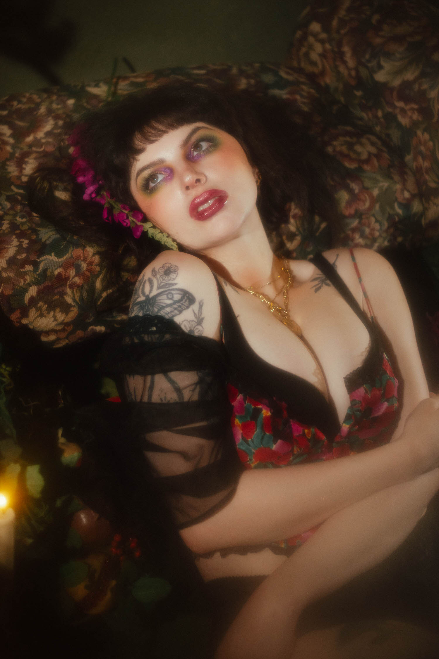 A woman poses in a red, pink, and green floral lingerie top with a sheer black vintage robe draped over one arm, against a floral tapestry backdrop with pink flowers tucked into her hair in this vintage lingerie boudoir photoshoot, Royal Lune Photo.