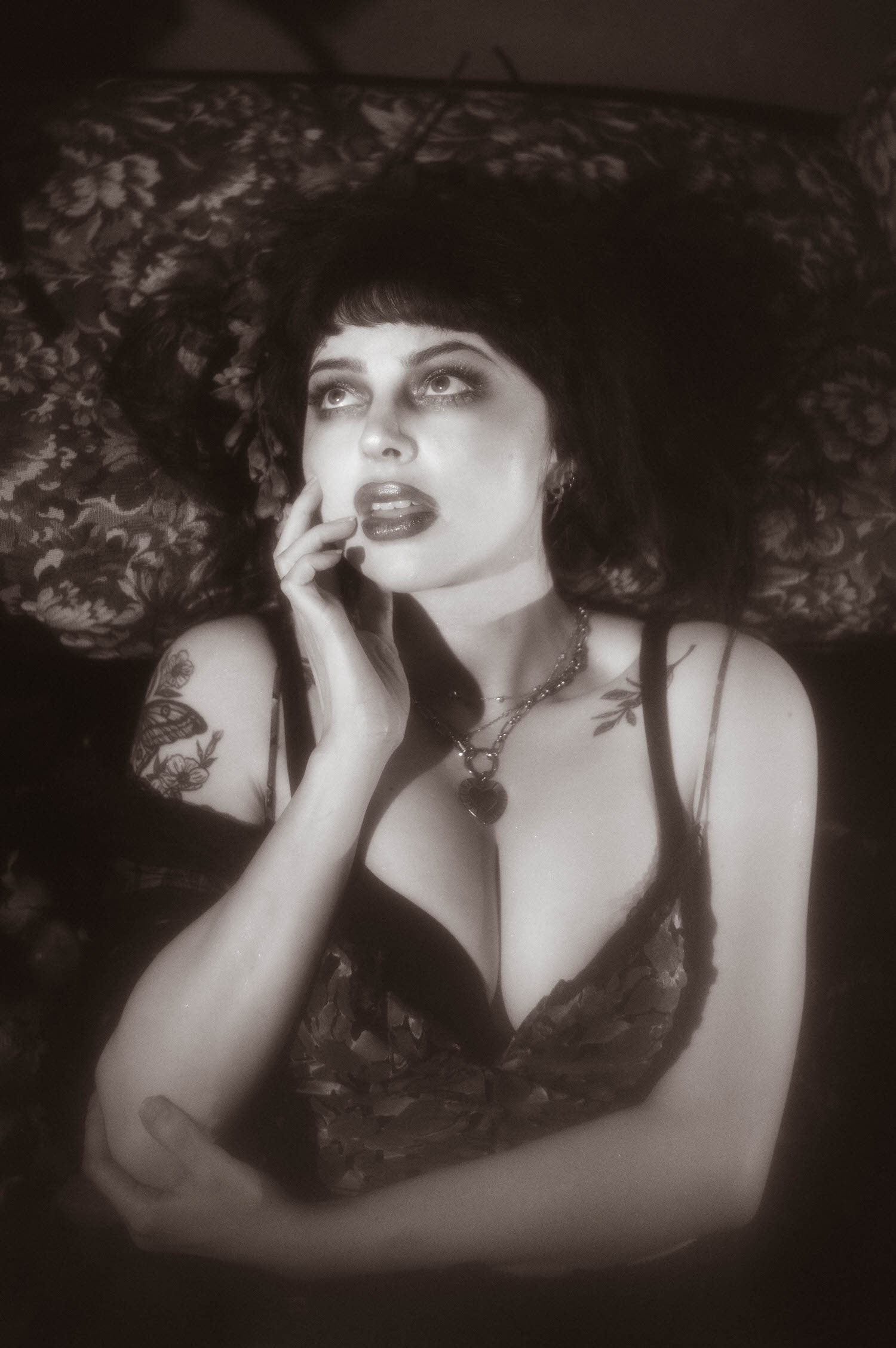 A black and white portrait shows a woman in a sheer black vintage robe and lace trimmed lingerie top resting her chin on her hand against a floral tapestry backdrop in this vintage lingerie boudoir photoshoot, Royal Lune Photo.