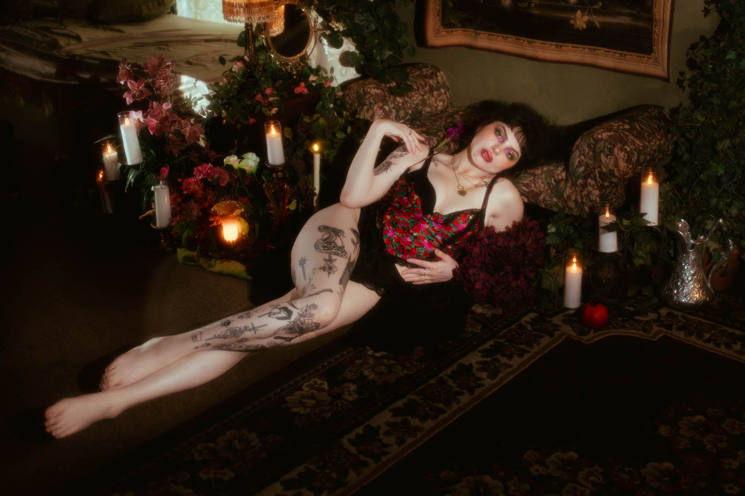 A woman reclines with her legs raised on a vintage tapestry sofa, wearing a sheer black vintage robe over a red, pink, and green floral lingerie set, surrounded by flameless candles and an antique silver pitcher in this red rose boudoir photoshoot at Royal Lune Studio, Royal Lune Photo.