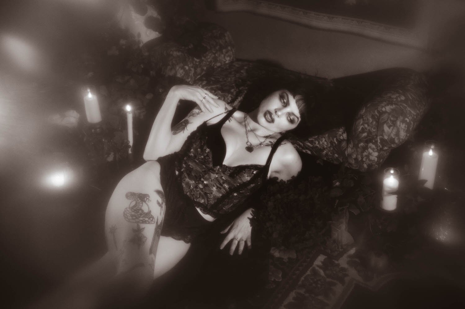 A black and white portrait shows a woman reclining on a tapestry sofa in a sheer black vintage robe, with visible thigh and hand tattoos and a layered necklace, in this dark romantic boudoir photography session, Royal Lune Photo.