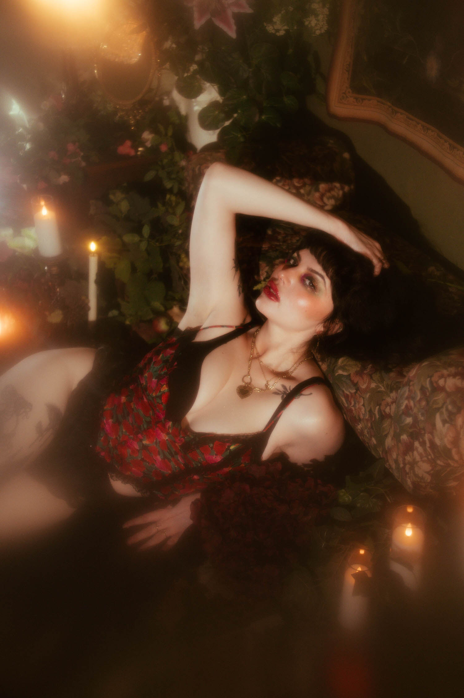 A woman lies back with one arm raised above her head, wearing a sheer black vintage robe over a red, pink, and green floral lingerie set and layered gold necklaces, lit by candlelight against a floral tapestry backdrop in this vintage lingerie boudoir photoshoot, Royal Lune Photo.