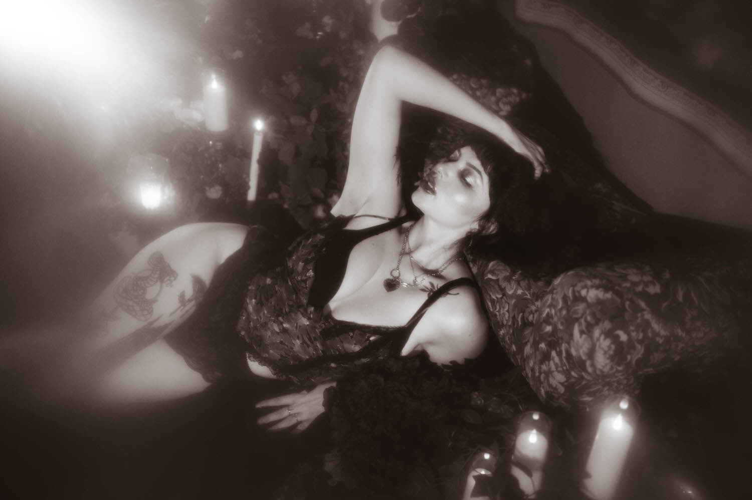 A black and white portrait shows a woman reclining in a sheer black vintage robe with her arm raised, wearing a lace trimmed lingerie set and layered necklaces beside lit candles in this fine art boudoir photography session, Royal Lune Photo.