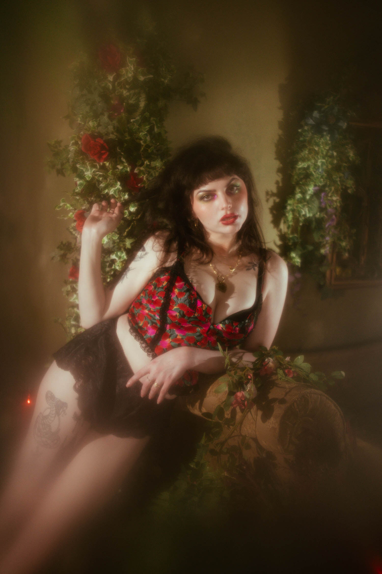 A woman sits leaning forward in a red, pink, and green floral lingerie set with black lace trim, styled beside an antique mirror and a wall of ivy and red roses in this fine art boudoir photography session, Royal Lune Photo.