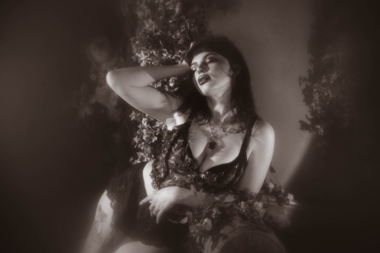 A black and white portrait shows a woman leaning back against a wall of climbing greenery, wearing a layered pendant necklace and lace lingerie in this boudoir photoshoot ideas inspired session, Royal Lune Photo.