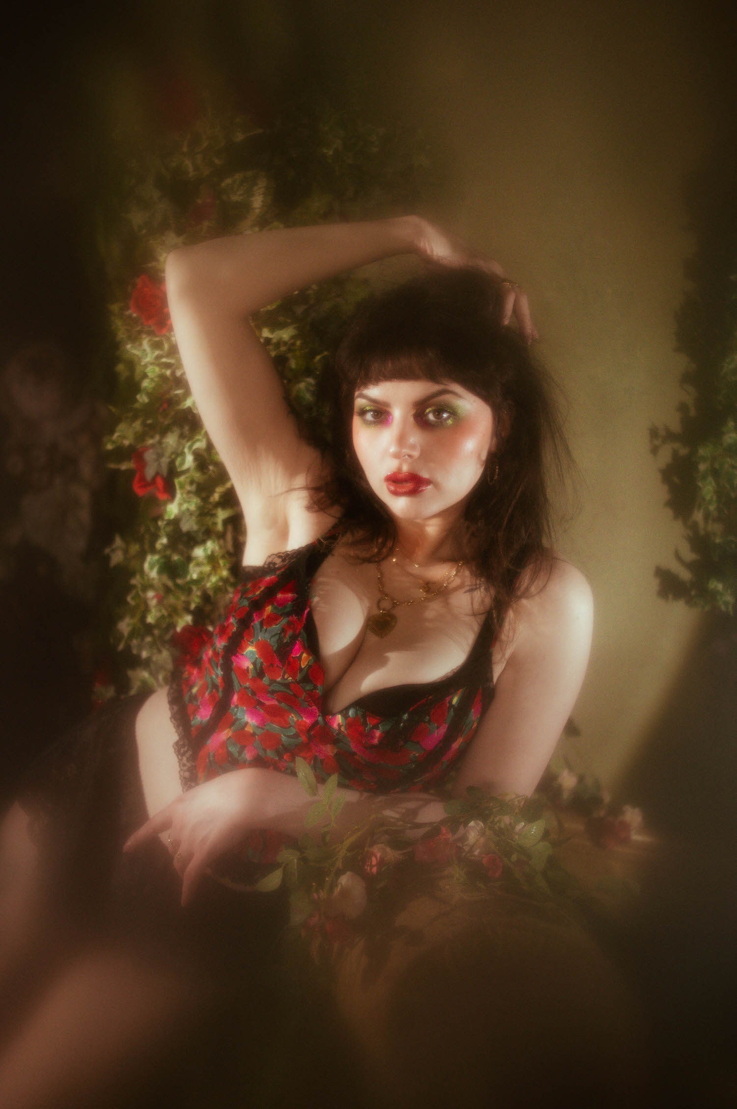 A woman poses with one arm raised above her head, wearing a red, pink, and green floral lingerie top against a wall of climbing ivy and red roses in this red rose boudoir photoshoot, Royal Lune Photo.