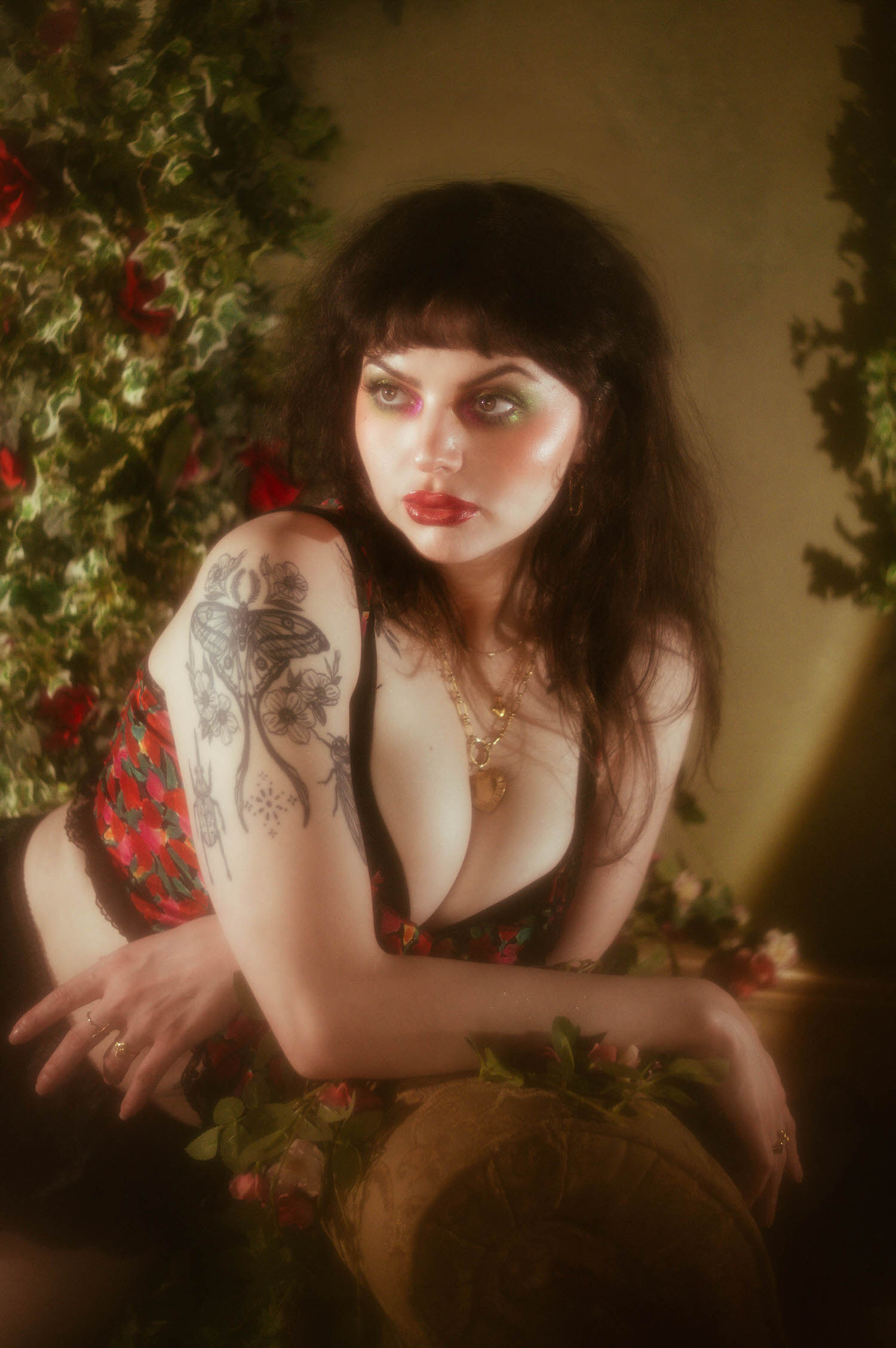 A close up portrait shows a woman with a floral and moth tattoo on her shoulder, wearing a red, pink, and green floral lingerie top and layered gold necklaces beside climbing ivy and red roses, Royal Lune Photo.