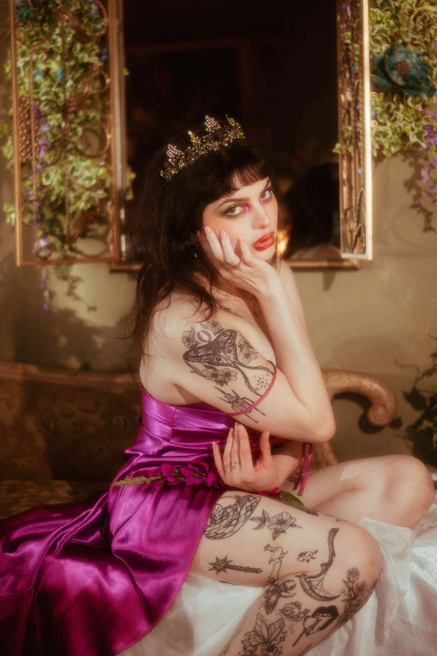 A bride wearing a magenta satin slip dress and a gold jeweled tiara rests her hand against her cheek during a bridal boudoir photography session, showing fine line moth and floral tattoos along her arm and leg inside Royal Lune Studio, Royal Lune Photo.