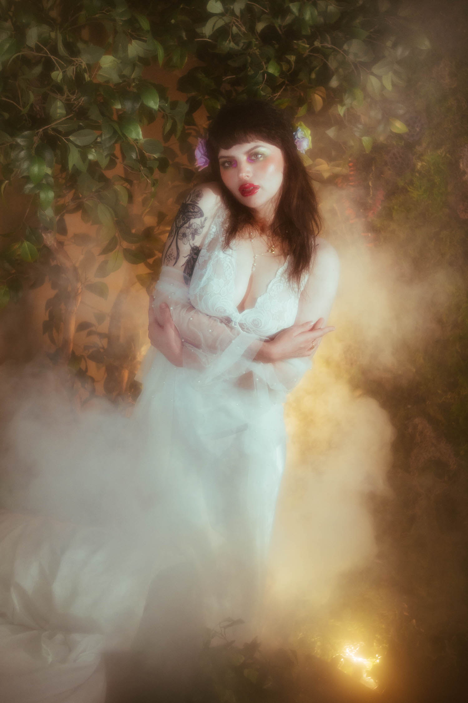 A bride wearing a vintage nightgown boudoir look stands with her arms crossed over her chest, a lilac rose tucked into her hair and soft fog drifting around her against a backdrop of greenery, Royal Lune Photo.