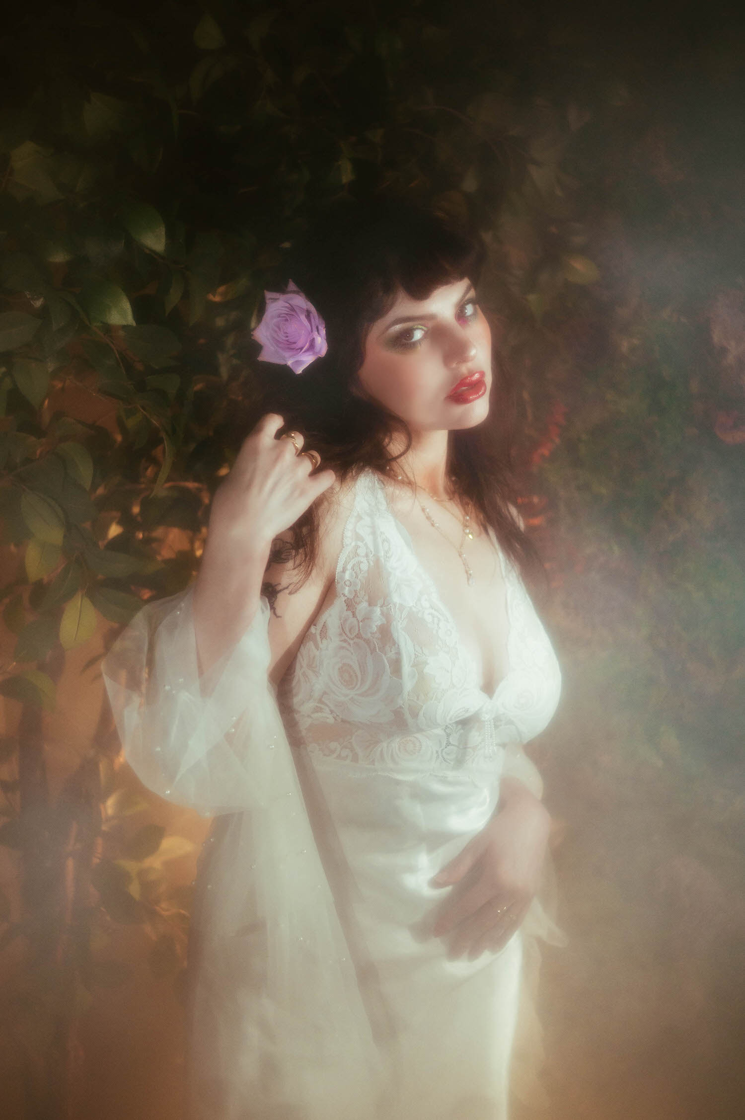 A bride in a vintage nightgown boudoir look glances over her shoulder, her sheer bell sleeve catching warm light as a lilac rose rests in her dark hair against a foliage backdrop, moody boudoir lighting shaping the soft shadows around her, Royal Lune Photo.