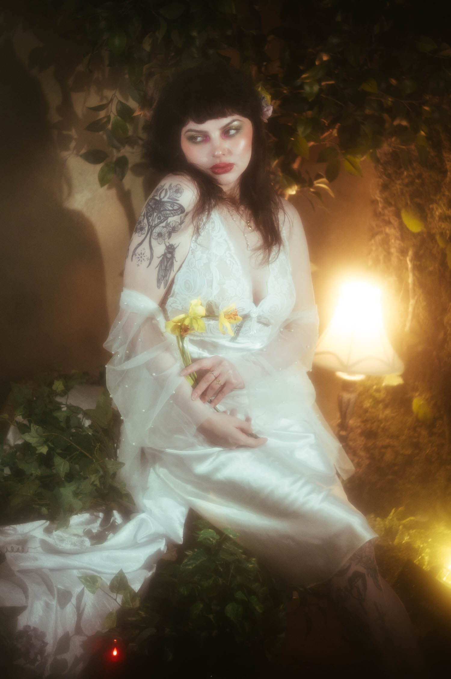 A bride sits in a vintage nightgown boudoir look holding yellow flowers, her tattooed arm visible as warm lamplight glows beside her during this bridal boudoir photography session at Royal Lune Studio, Royal Lune Photo.