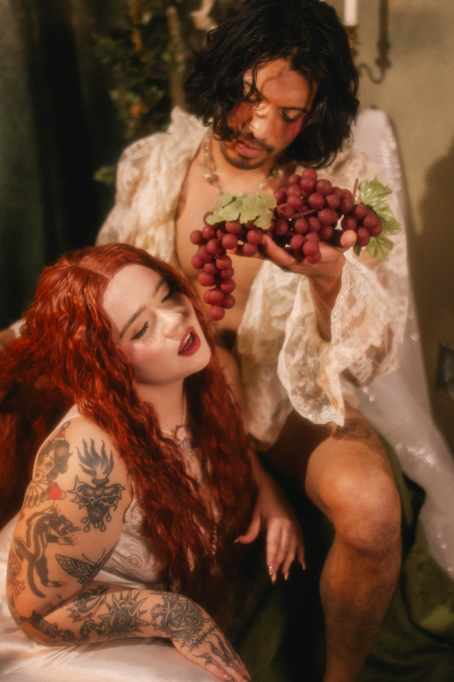 A couples boudoir photoshoot idea styled after a classical still life, the man holding a cluster of grapes above his partner as she leans back with eyes closed. Royal Lune Photo