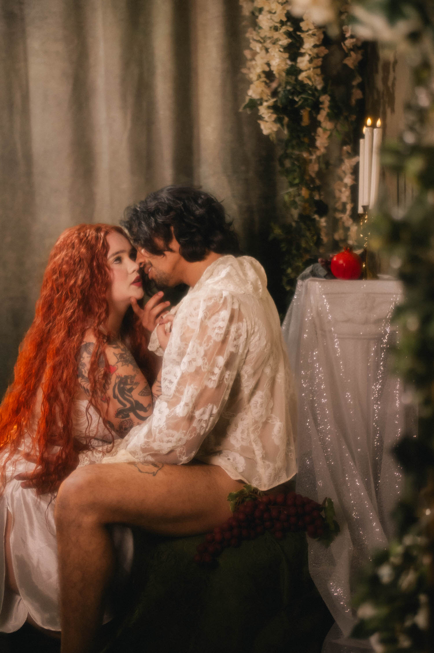 Dallas Fort Worth boudoir photographer captures an intimate exchange beside a candlelit pedestal table, a single pomegranate and trailing florals setting the scene. Royal Lune Photo