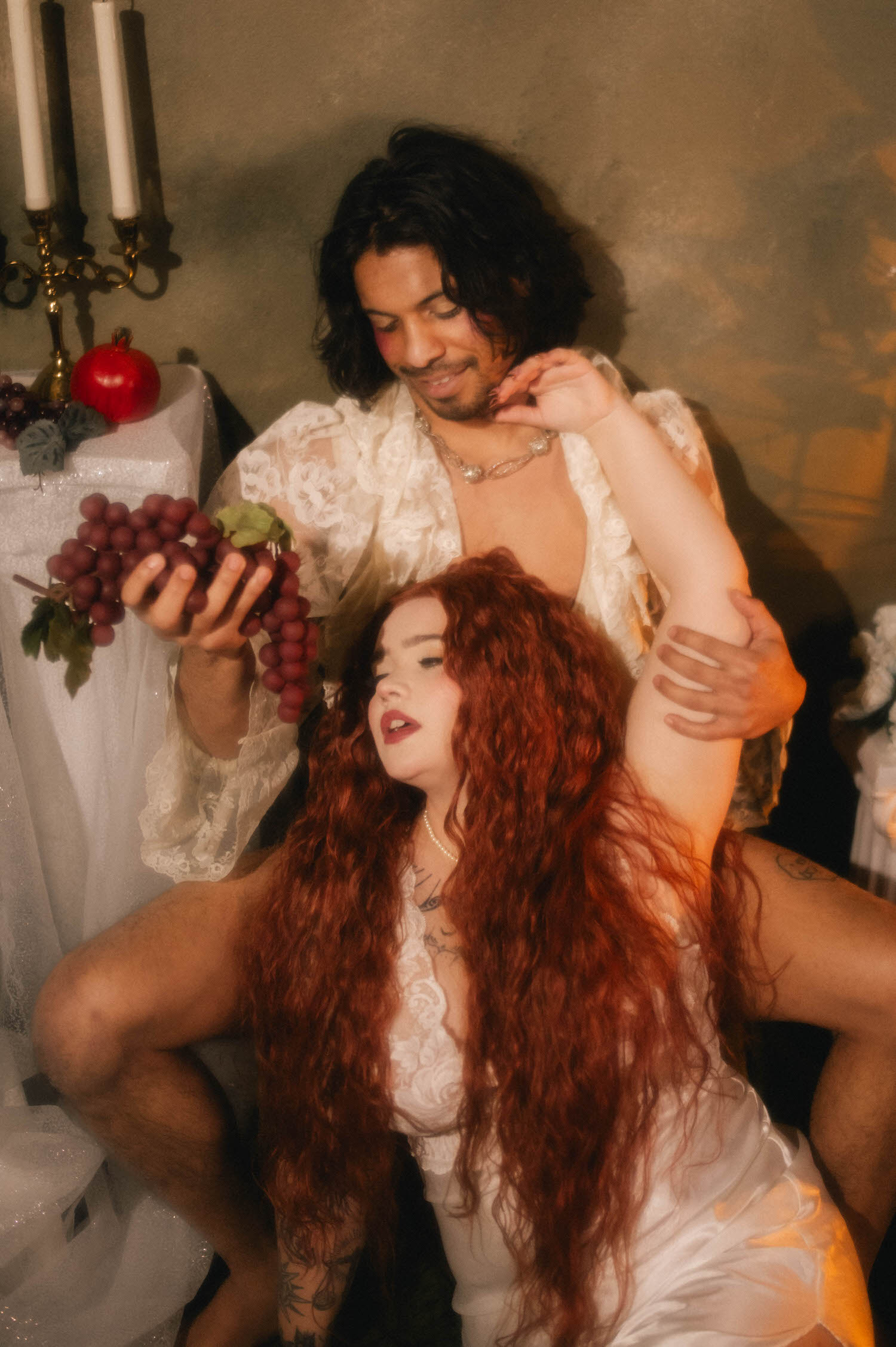 Renaissance inspired photoshoot detail shot of a couple surrounded by grapes and a pomegranate on a draped pedestal, the woman's head tilted back against her partner's arm as he feeds her grapes. Taken by Royal Lune Photo