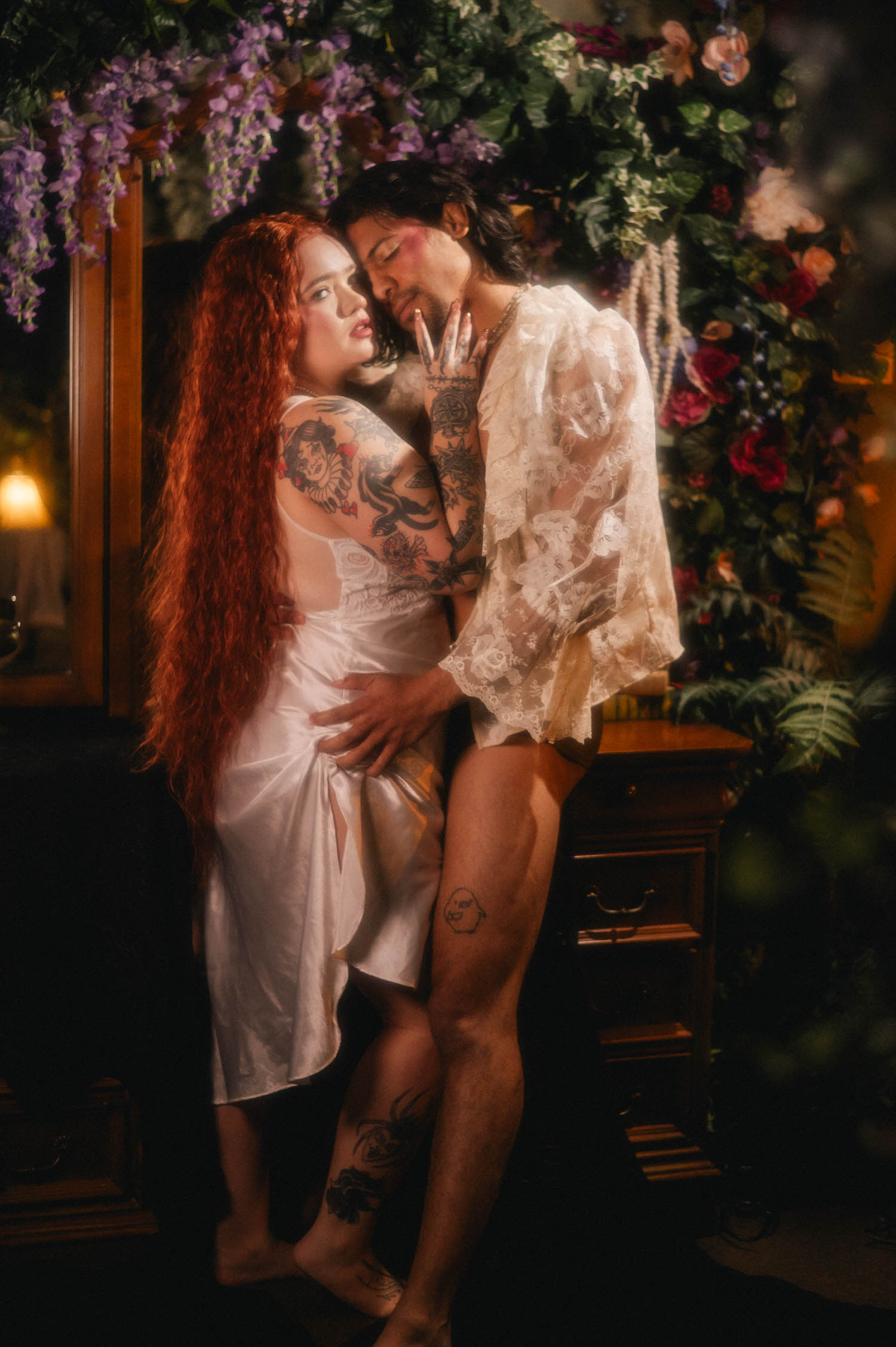 A queer couples boudoir moment captured beside a vintage wooden dresser, the woman glancing back over her shoulder as her partner embraces her from behind. Royal Lune Photo