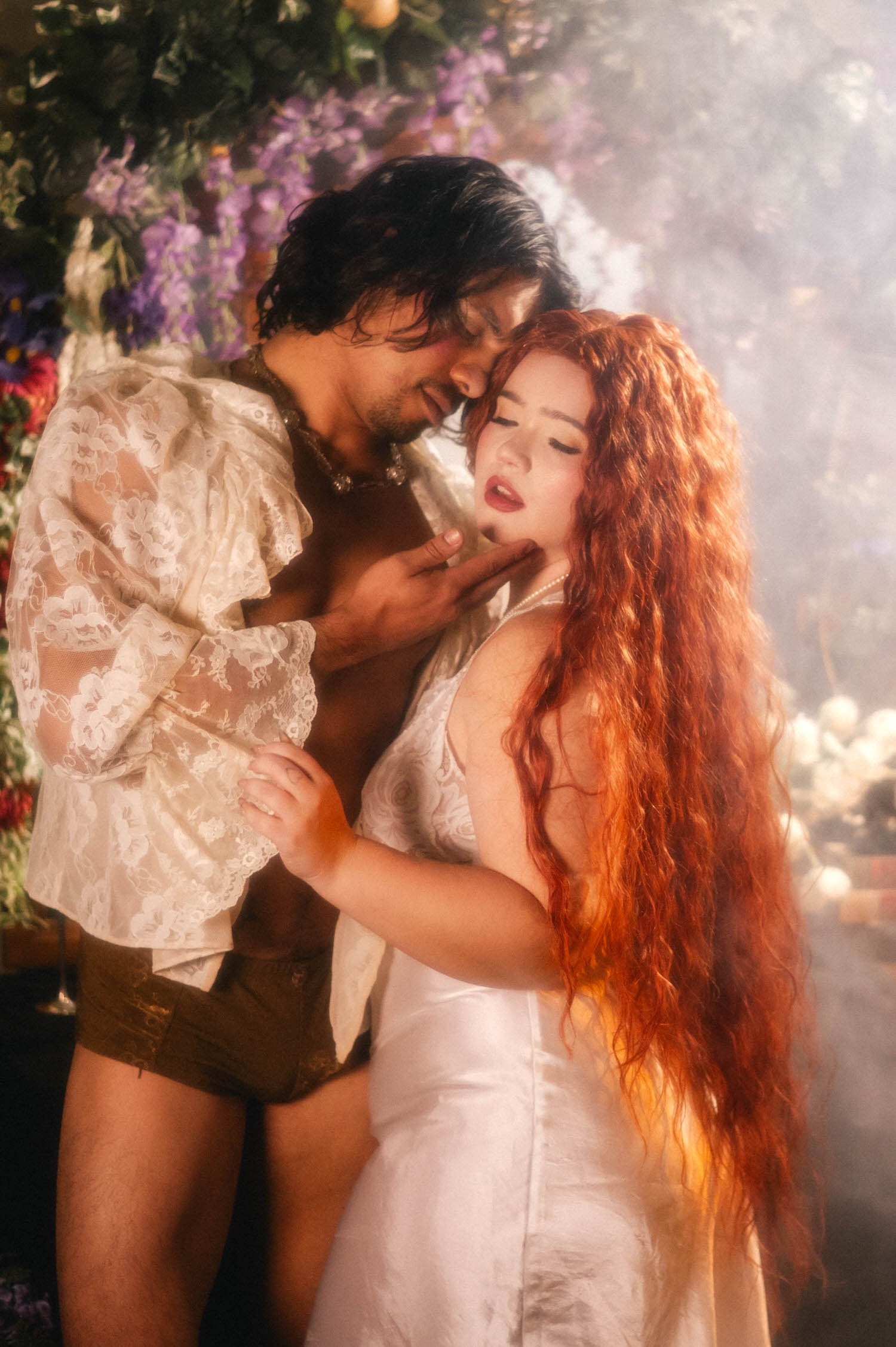 A Renaissance boudoir photoshoot embrace captured in soft fog, the man's hand resting at his partner's throat as florals glow behind them. Royal Lune Photo