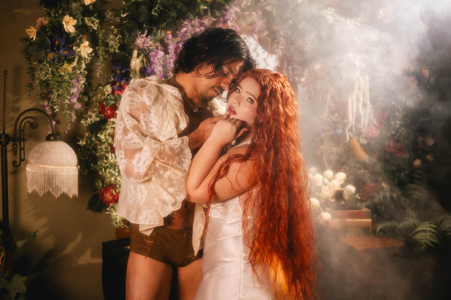 Romantic couples boudoir portrait inspired by Pre-Raphaelite art taken beside a vintage lamp and dense floral backdrop, the woman gazing directly into camera as her partner nuzzles close. Royal Lune Photo