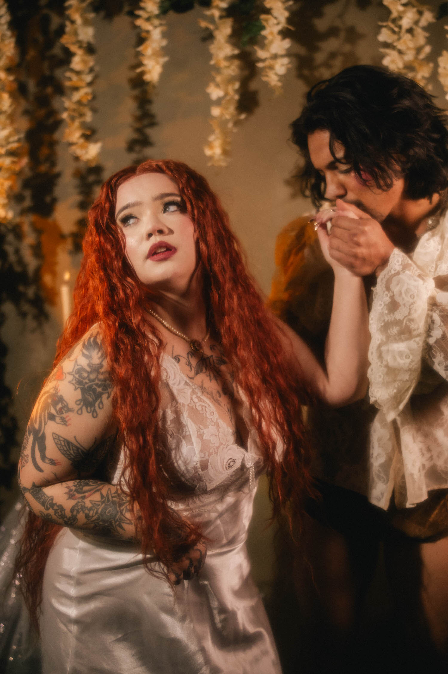 A bridal boudoir photoshoot moment shows a woman in a white lace slip watching her partner kiss her hand, hanging wisteria blooms framing the couple in warm light. Royal Lune Photo