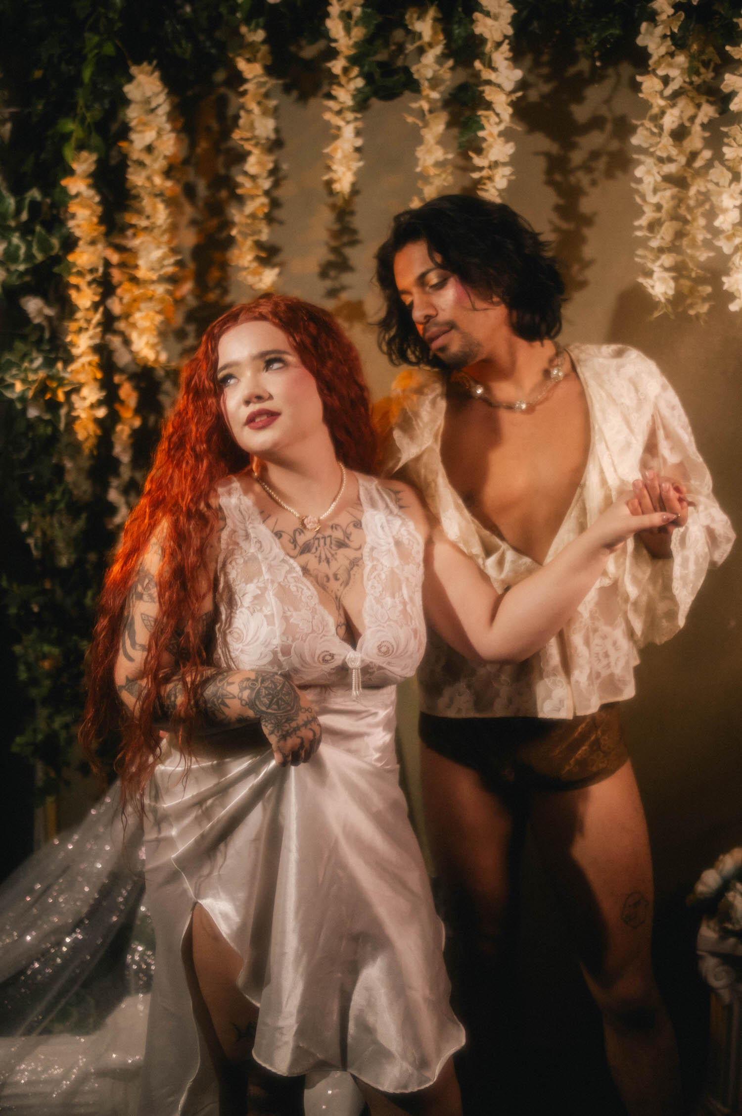 Couples boudoir photoshoot ideas come to life as a woman in a silky white gown stands beside her partner in a sheer lace robe, both barefoot beneath cascading white florals. Royal Lune Photo