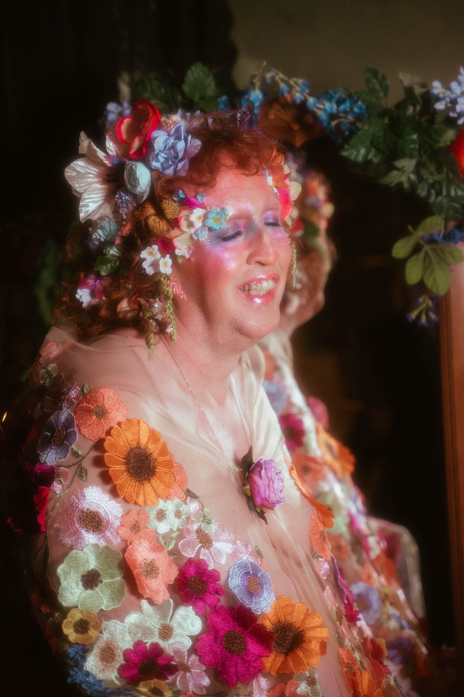 This LGBTQ+ photographer Dallas session captures a wide smile framed by a flower crown of white, blue, and coral blooms, with a rose pendant necklace resting against a sheer floral gown. Royal Lune Photo