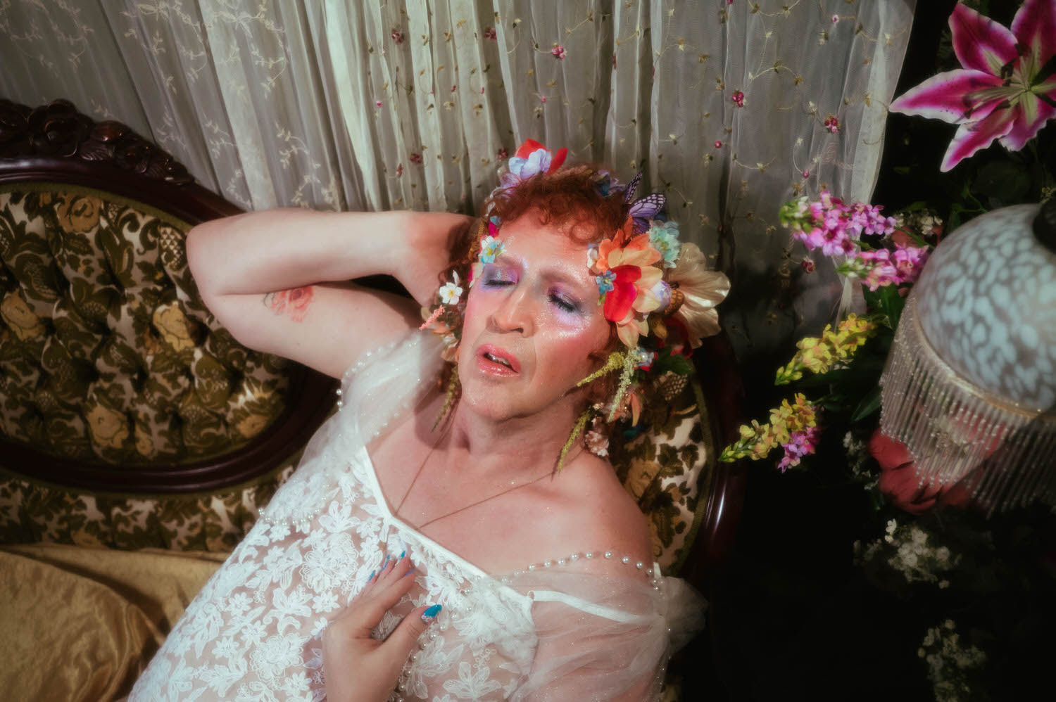 This trans boudoir photoshoot image shows a woman reclining on a tufted vintage sofa in a sheer white lace slip dress with pearl beaded straps, her flower crown resting against embroidered sheer curtains and a stargazer lily nearby. Royal Lune Photo