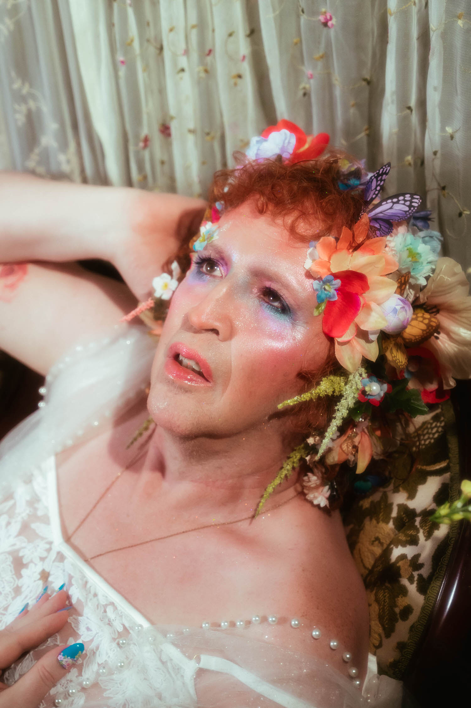 A close up from this LGBTQ+ photographer Dallas session shows a woman gazing upward, her flower crown of red, peach, and blue blooms framing glittered makeup above a pearl trimmed white lace neckline. Royal Lune Photo