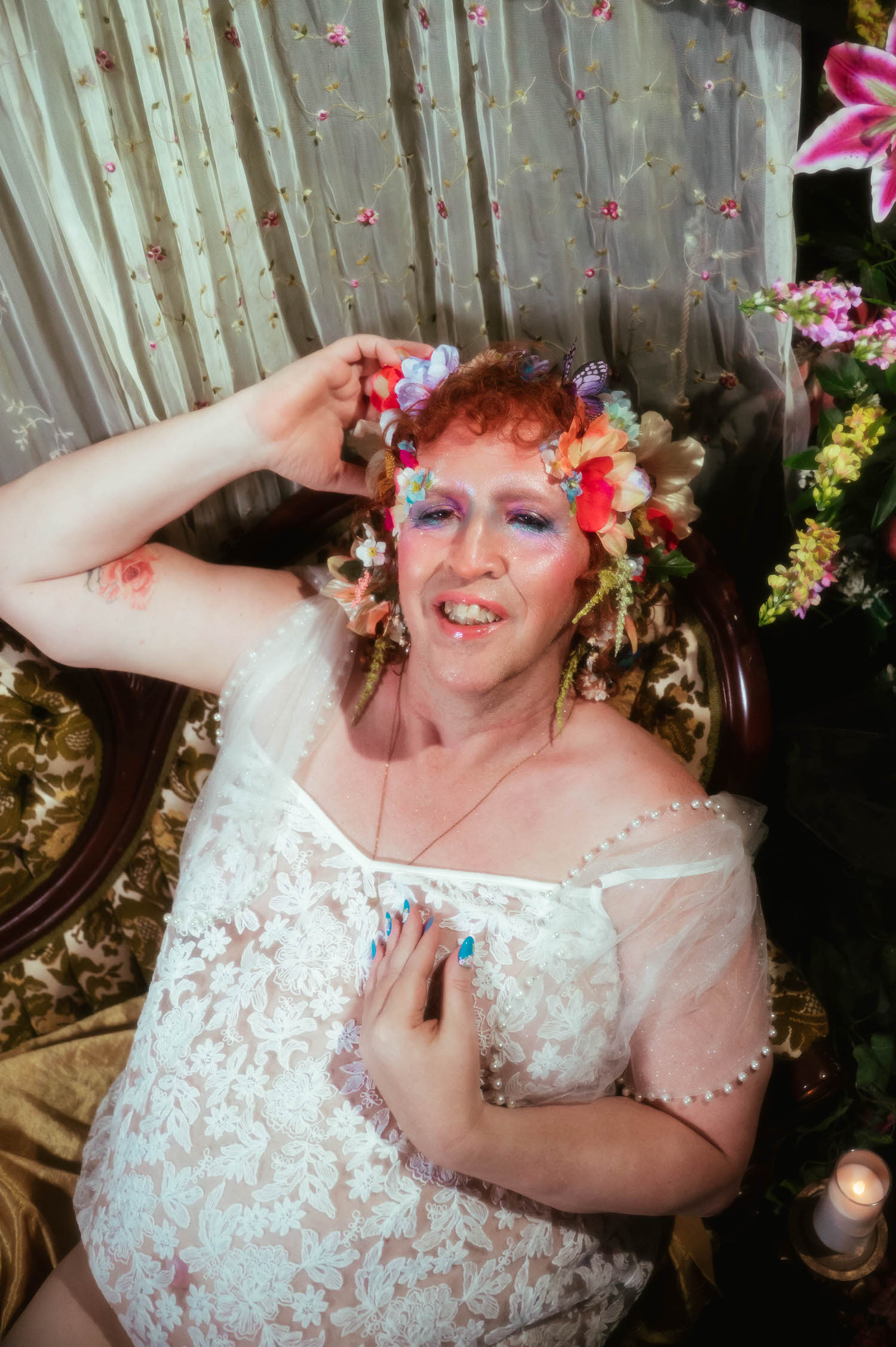 This self expression through art image shows a woman smiling softly with her hand adjusting her flower crown, a rose tattoo visible on her raised arm, wearing a sheer white lace slip dress with pearl beading. Royal Lune Photo