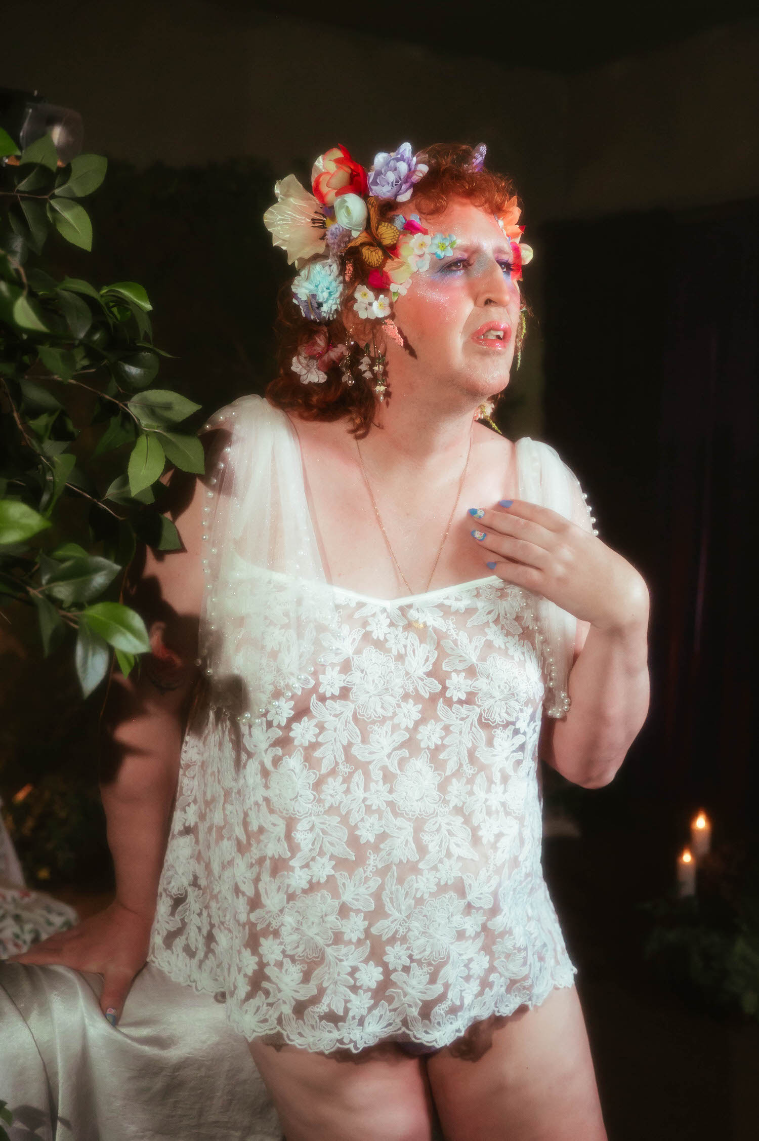 In this trans boudoir photoshoot, a woman stands beside greenery in a sheer white lace top with pearl trimmed sleeves, one hand resting at her chest as she looks down thoughtfully. Royal Lune Photo