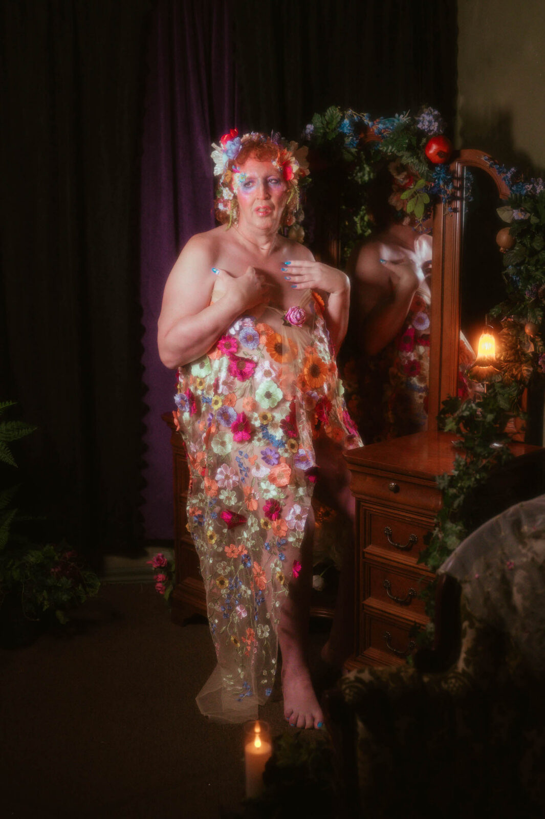 A trans boudoir photoshoot at Royal Lune Studio in Denton, TX, shows a woman standing before an antique mirror wrapped in ivy and pomegranates, wearing a sheer floral embroidered gown in shades of coral, lavender, and cream, with a lit candle glowing at her feet. Royal Lune Photo