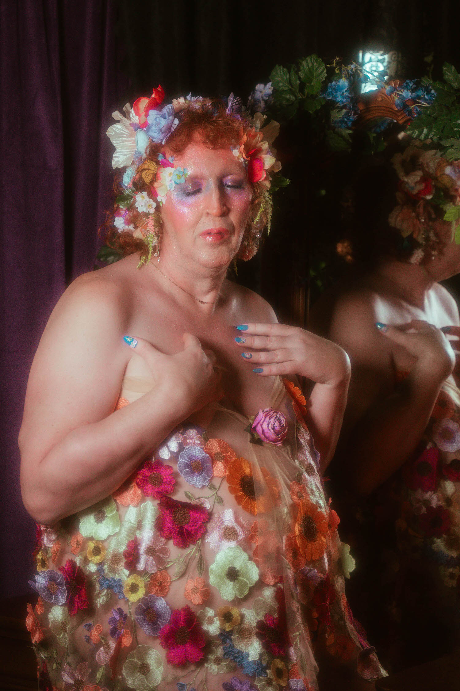 This gender affirming boudoir portrait captures a woman with closed eyes and a flower crown of red, lavender, and cream blooms, her hand resting over a sheer floral embroidered gown at Royal Lune Studio. Royal Lune Photo