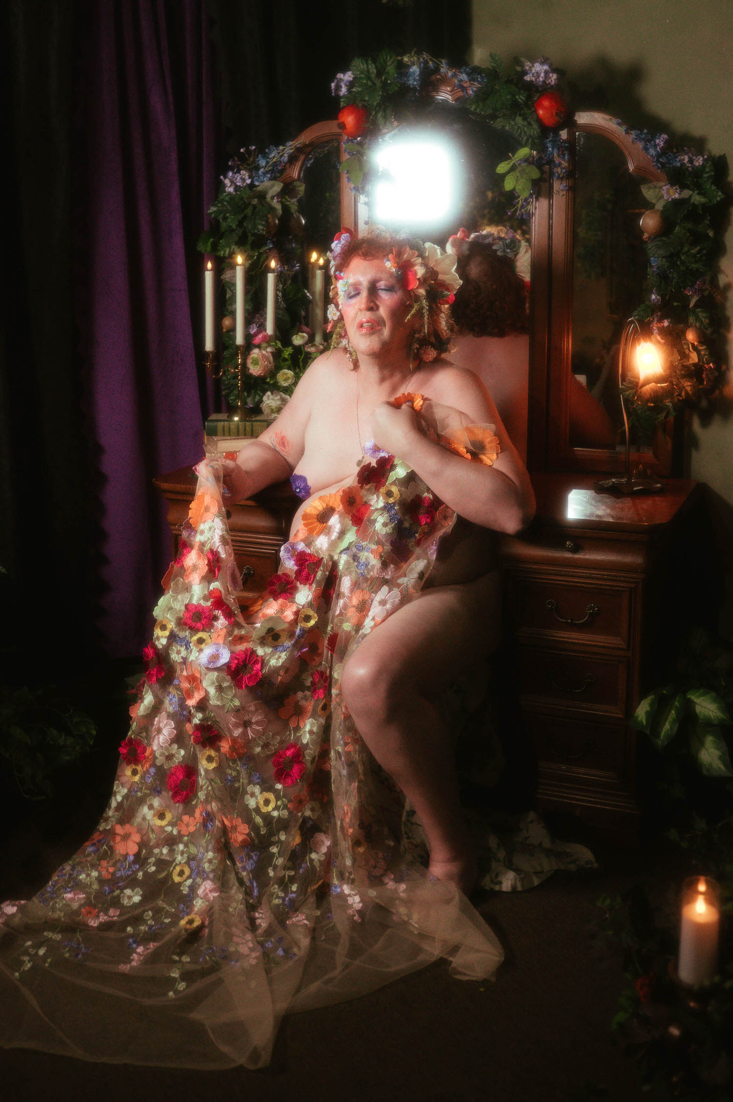 In this trans boudoir photoshoot, a woman sits at an antique vanity lit by a candelabra, her floral embroidered gown pooling around her as she gathers the sheer fabric at her chest. Royal Lune Photo