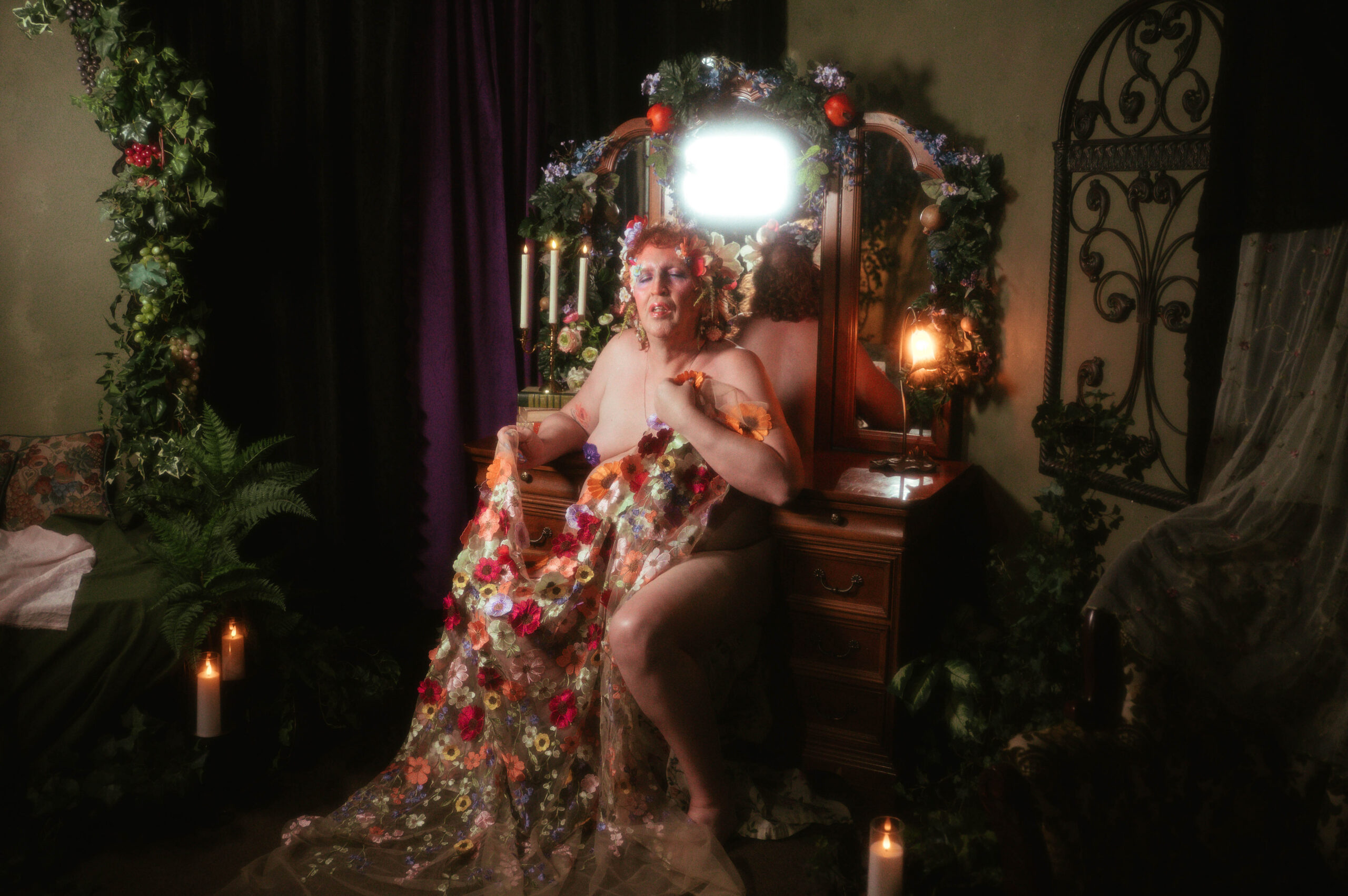 A wide shot from this trans boudoir photoshoot in Denton, TX, reveals the full studio setting, with an ivy and grape draped mirror, wrought iron wall decor, and a floral embroidered gown pooling across the floor beside lit candles. Royal Lune Photo