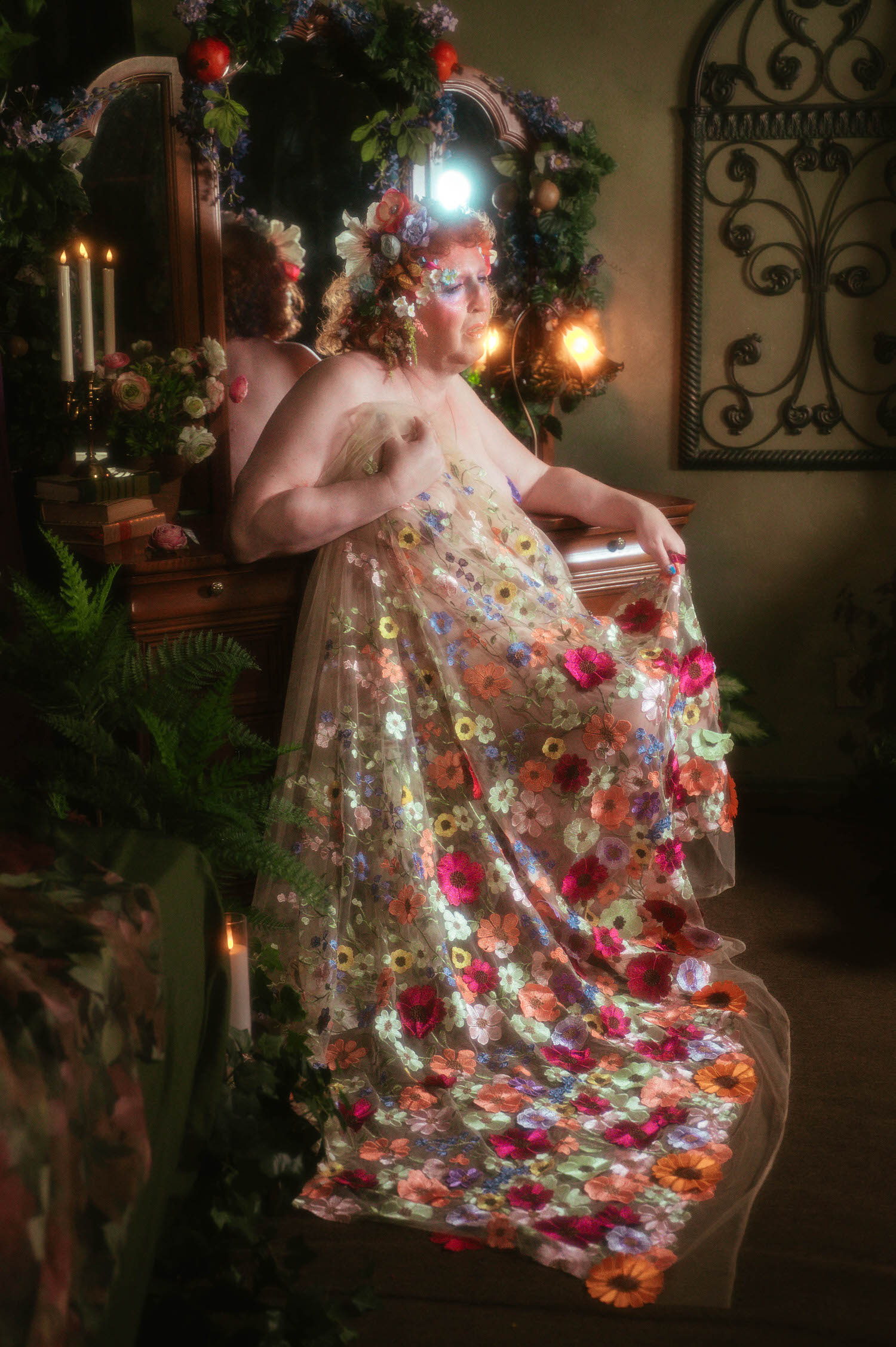 This gender affirming boudoir photo shows a woman leaning against an antique vanity, her floral embroidered gown draped over one arm as candlelight and greenery fill the studio around her. Royal Lune Photo