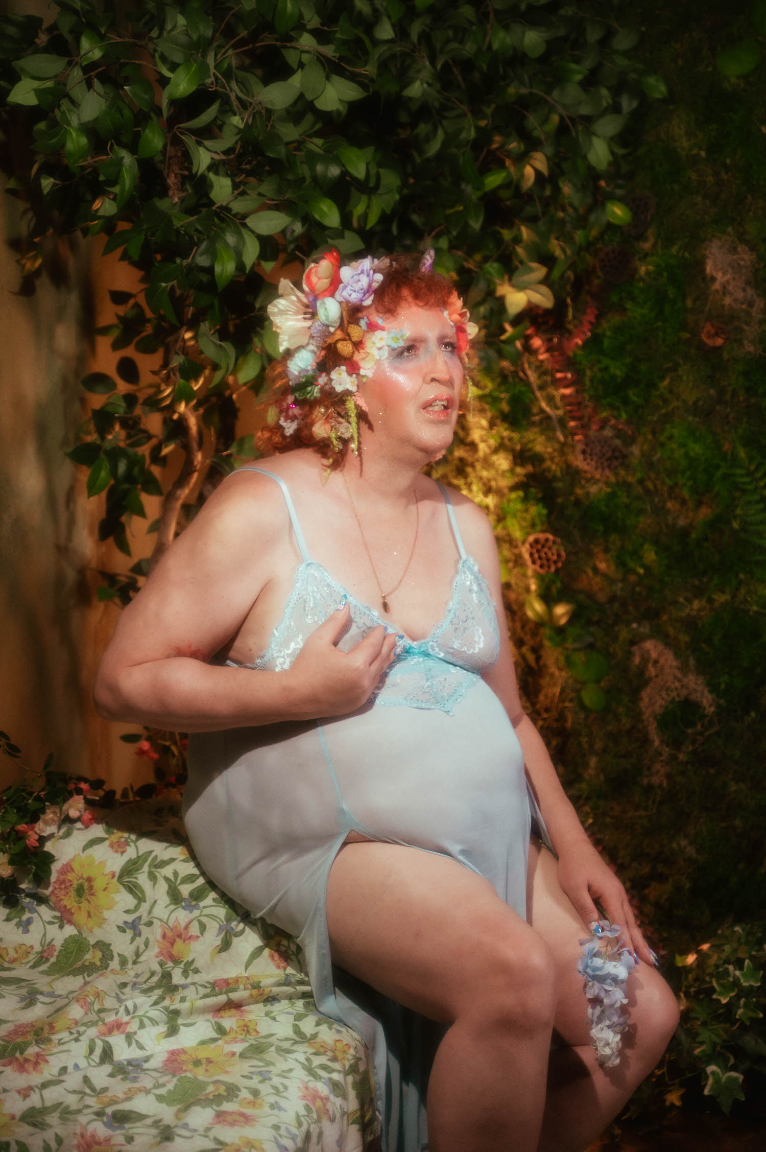 In this trans boudoir photoshoot moment, a woman sits in profile against a backdrop of trailing ivy, wearing a pale blue lace trimmed slip dress with a matching floral garter wrapped around her thigh. Royal Lune Photo