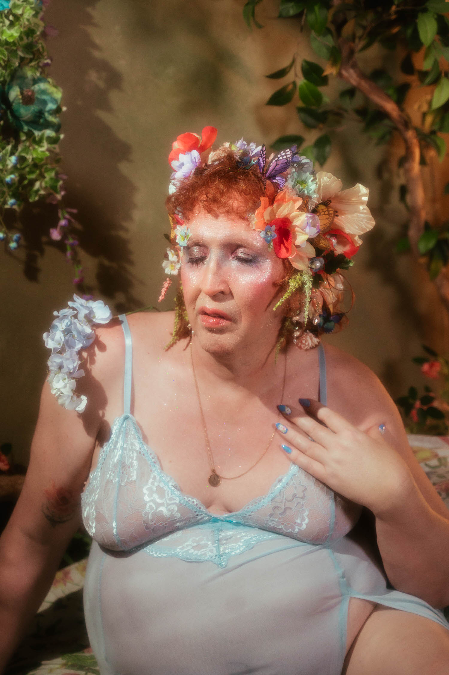 This gender affirming boudoir portrait shows a close up of a woman with closed eyes, a flower crown of red poppies and butterflies framing her face above a pale blue lace bodice and a gold pendant necklace. Royal Lune Photo