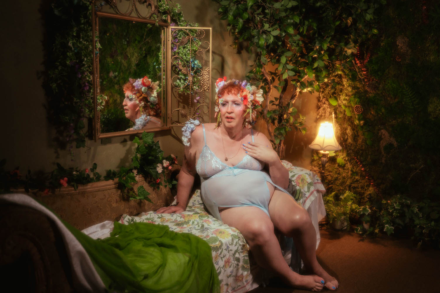This LGBTQ+ photographer Dallas session captures a woman seated on a floral chaise in a greenery filled corner of Royal Lune Studio, wearing a pale blue lace trimmed slip dress with a flower crown and matching floral garter, warm lamp light glowing beside her. Royal Lune Photo