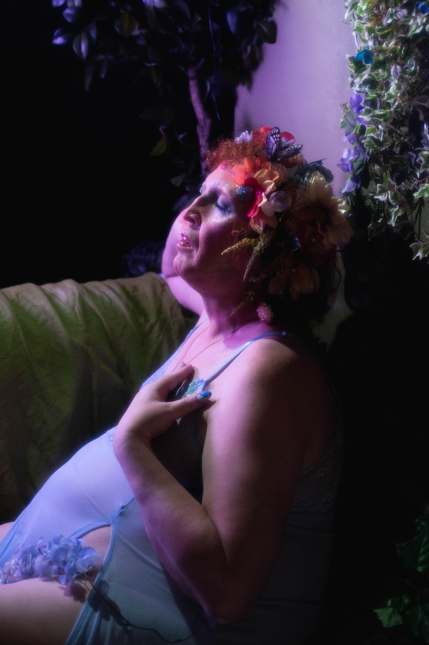 This gender affirming boudoir portrait shows a woman in profile with her head tilted back under a purple toned lighting gel, her flower crown catching the light against a backdrop of trailing ivy at Royal Lune Studio. Royal Lune Photo