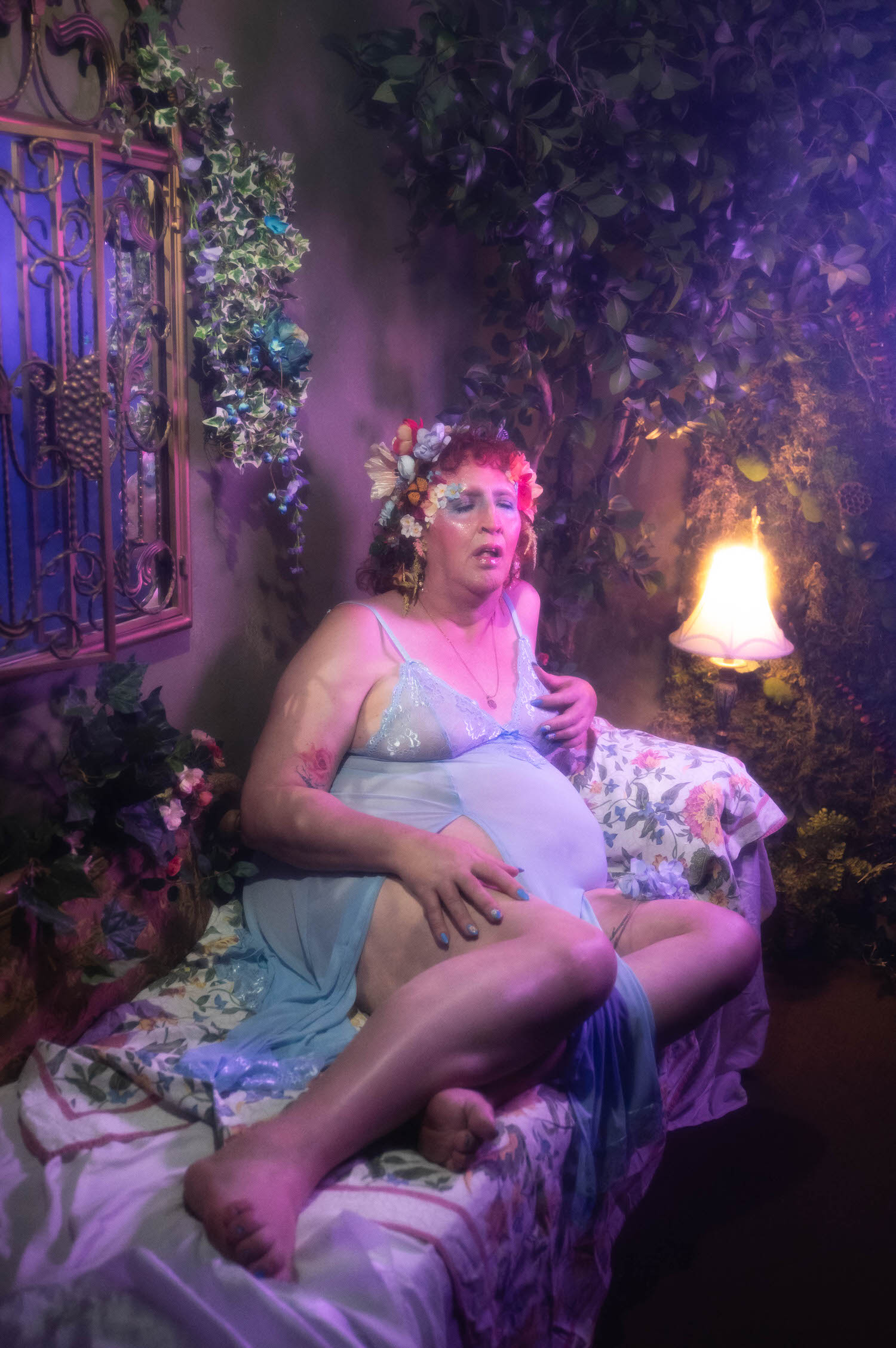 A gender affirming boudoir photo shows a woman reclining on a floral chaise under a soft purple lighting gel, her pale blue lace slip dress and floral garter visible as warm lamp light glows beside her. Royal Lune Photo