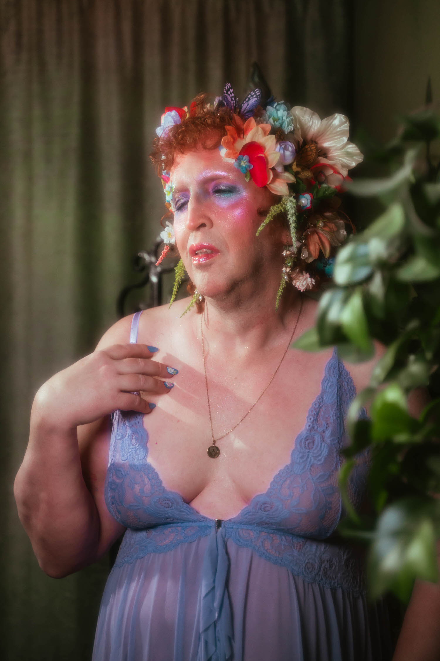 This gender affirming boudoir portrait shows a woman with closed eyes and glossy pink lips, her flower crown of red and cream blooms framing soft glittered makeup above a lavender lace bodice. Royal Lune Photo