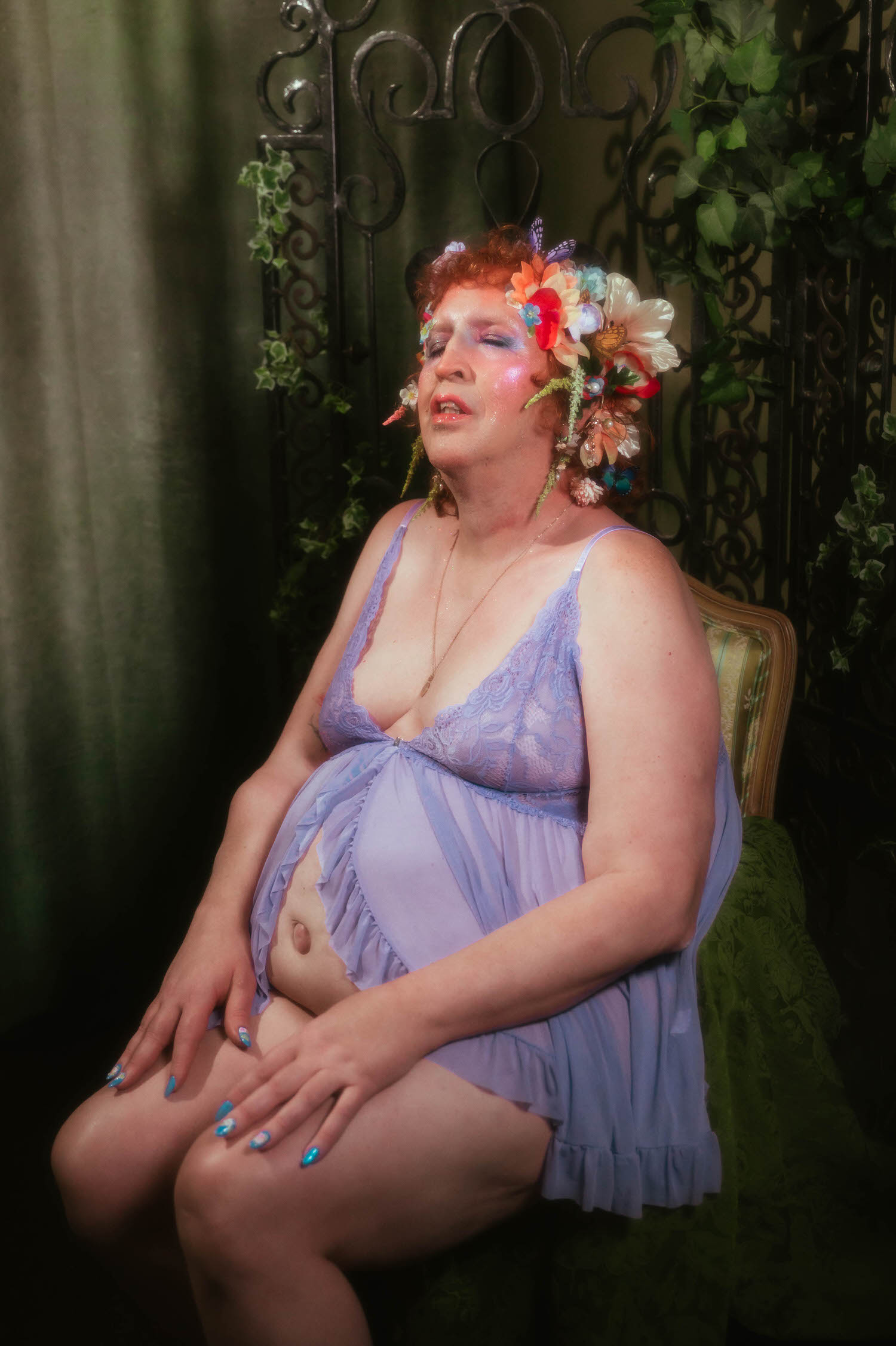 In this trans boudoir photoshoot, a woman sits in a vintage striped chair against a dark wrought iron screen woven with ivy, wearing a lavender lace trimmed babydoll with her hands resting on her knees. Royal Lune Photo