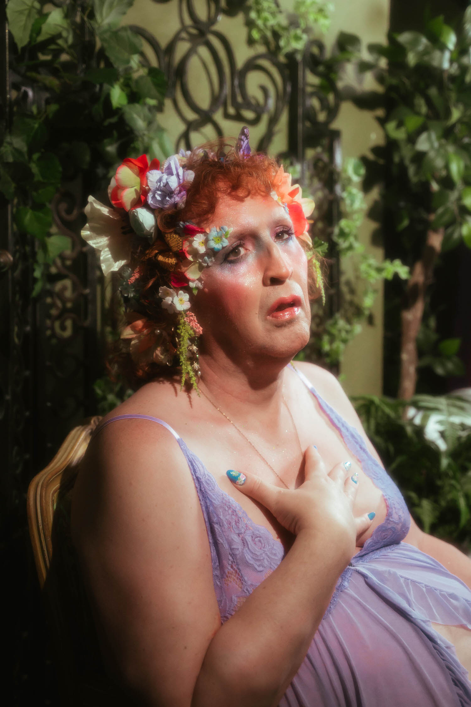 This LGBTQ+ photographer Dallas session captures a woman seated in front of greenery and an ornate iron screen, one hand resting at her chest over a lavender lace trimmed babydoll lingerie set. Royal Lune Photo