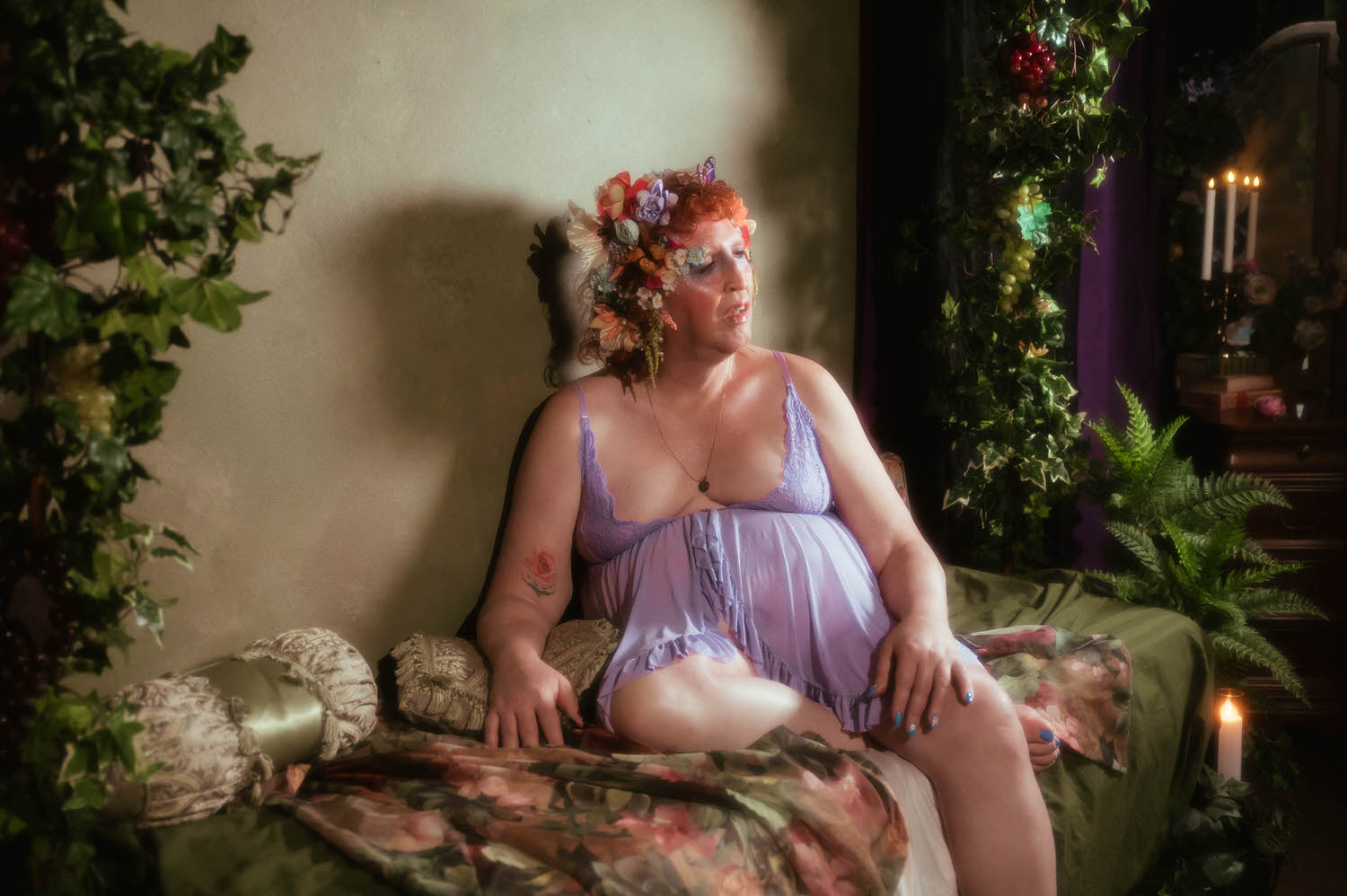 In this trans boudoir photoshoot, a woman sits on a green satin chaise beneath hanging grape clusters, wearing a lavender lace trimmed babydoll lingerie set, a small rose tattoo visible on her inner arm. Royal Lune Photo