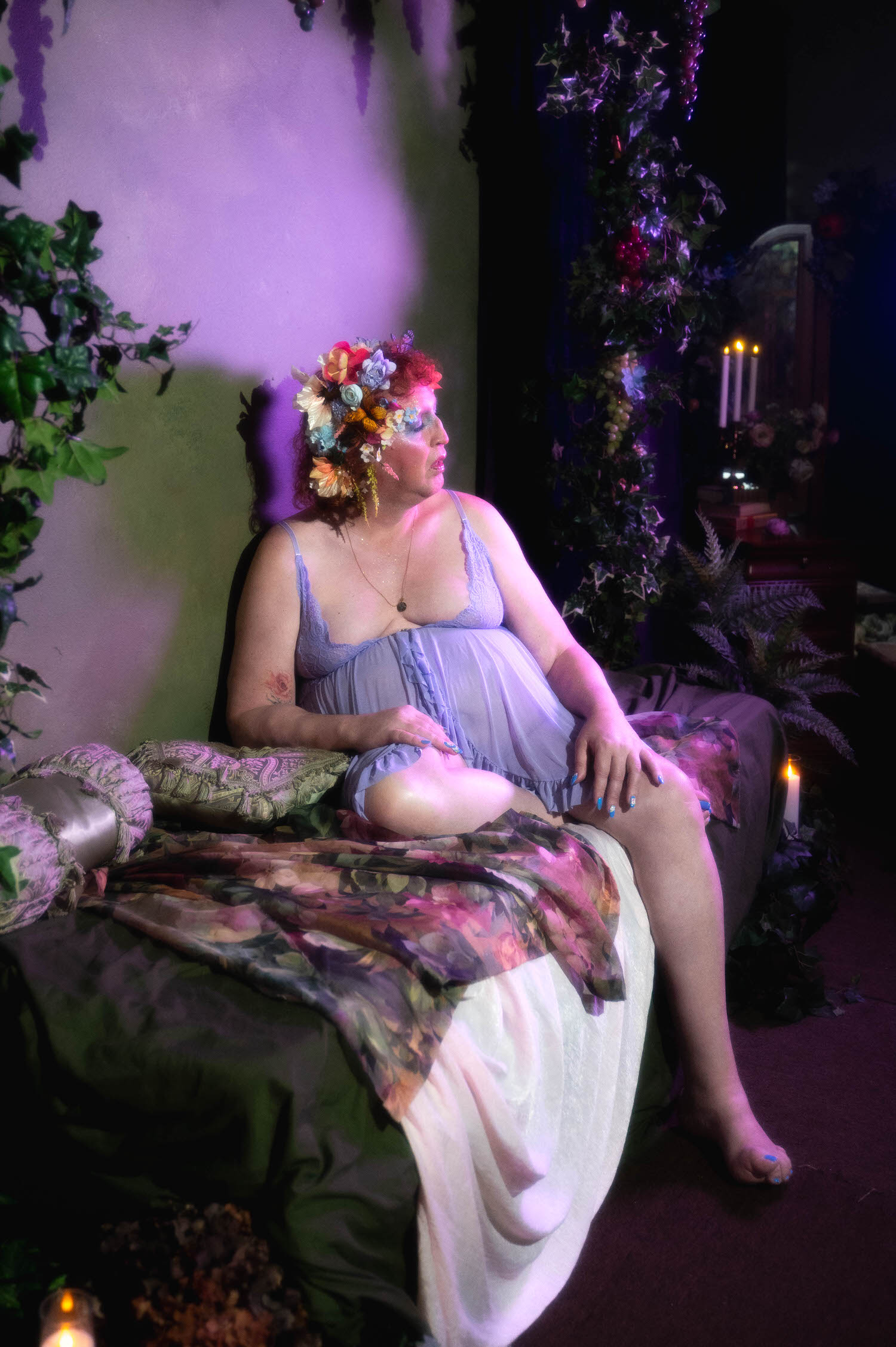 This LGBTQ+ photographer Dallas boudoir session captures a woman under soft purple lighting, seated on a chaise with a floral tapestry throw, a rose tattoo visible on her arm beside candlelight and trailing ivy. Royal Lune Photo