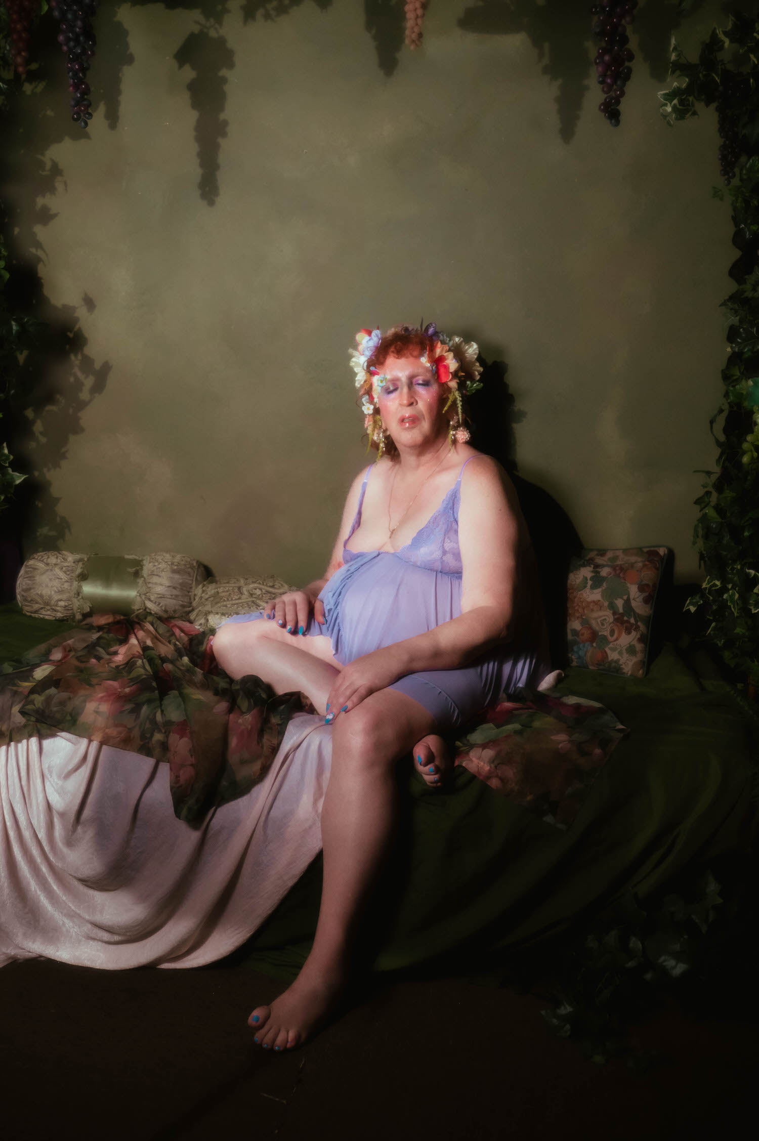 This gender affirming boudoir photo shows a wide shot of a woman seated cross legged on a chaise draped in a floral tapestry blanket, surrounded by ivy and hanging grape clusters against a sage green wall. Royal Lune Photo
