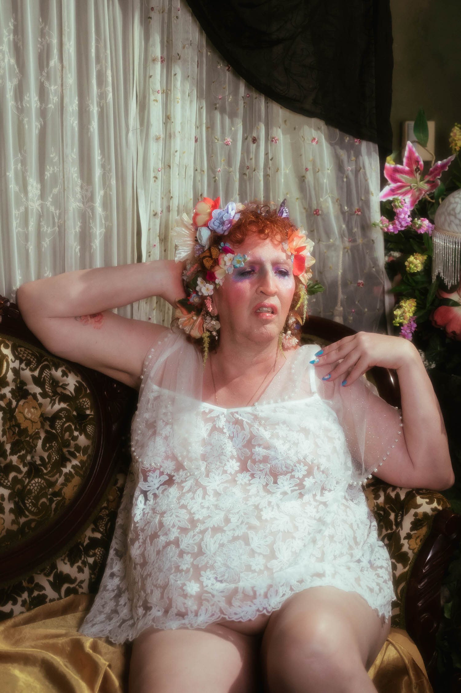 This gender affirming boudoir portrait captures a woman with one arm raised behind her head, a small rose tattoo visible on her inner arm, wearing a sheer white lace slip dress with pearl detailing. Royal Lune Photo