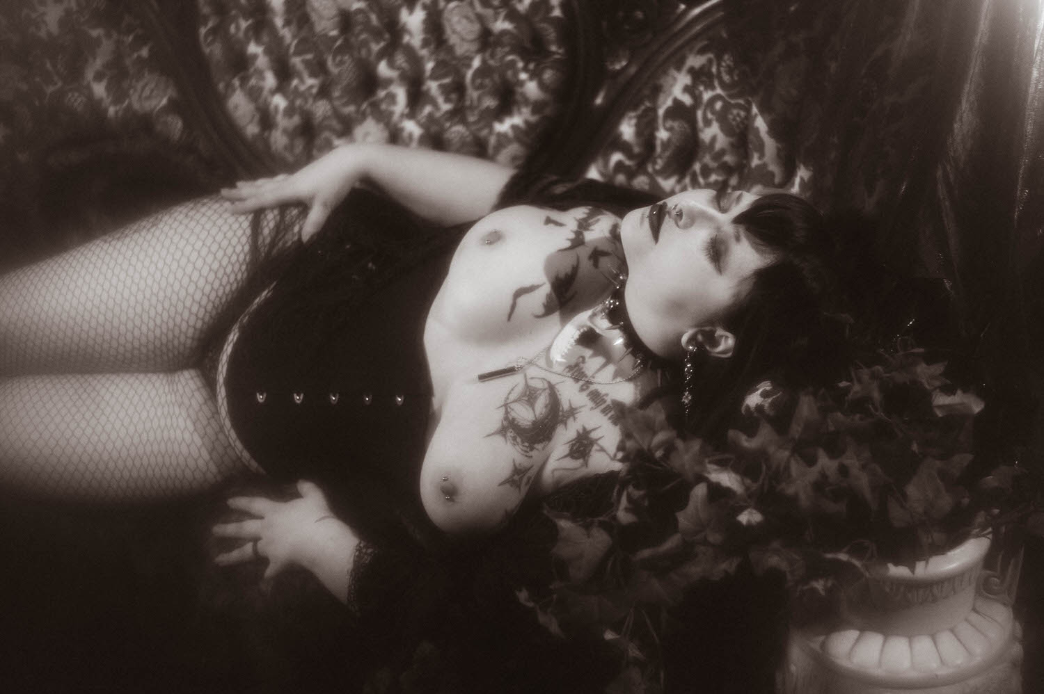 A black and white photo shows a bride reclining in a black corset and fishnet tights with a spiked choker, surrounded by ivy on a patterned Victorian sofa for this black corset boudoir photoshoot, Royal Lune Photo.