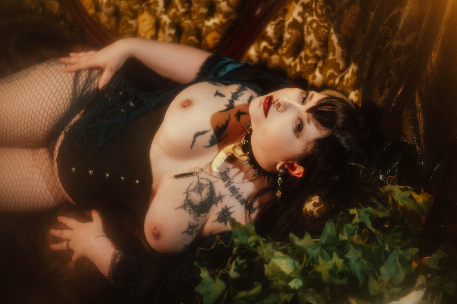 A bride reclines in a black corset and fishnet tights with a spiked choker and bone shaped pendant, sheer black fabric draped over a glowing lamp beside her for this gothic bridal boudoir session, Royal Lune Photo.